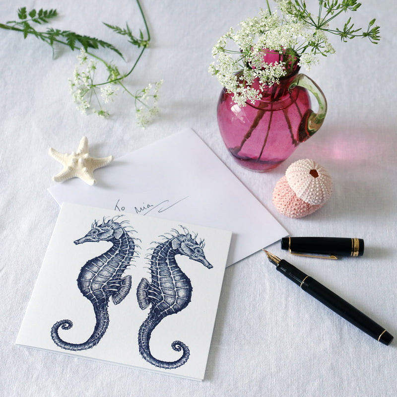 greeting card with illustrated navy seahorses back to back on a white background lying on a white table cloth with a fountain pen, hand written envelope shells and a small cranberry glass jug with wild flowers in 