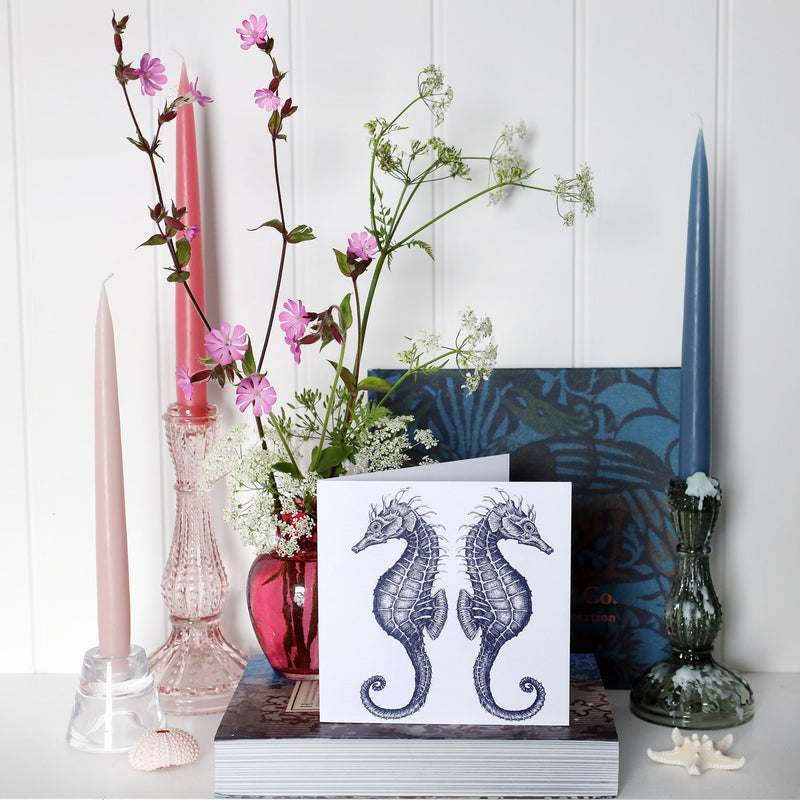 greeting card with illustrated navy seahorses back to back on a white background on shelf with pink and blue candles in candlesticks and a small cranberry glass jug with wild flowers in