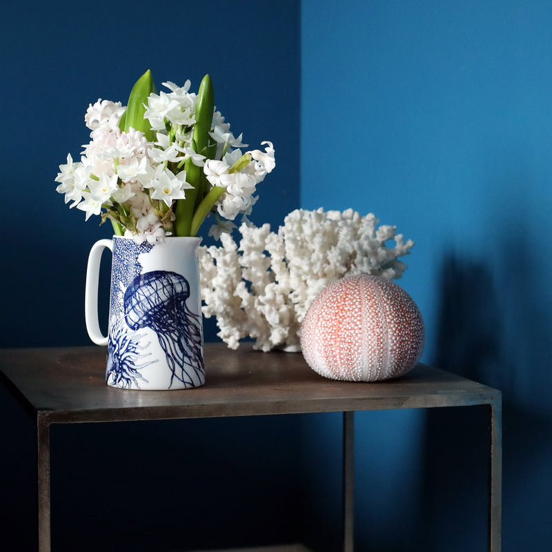 A white bone china jug with navy illustrations of a sea anemone, coral & jellyfish. There are soft pink hyacinths and pretty white daffodils in the jug and they are sitting on a metal table with some coral and a sea urchin, all against a dark teal wall.