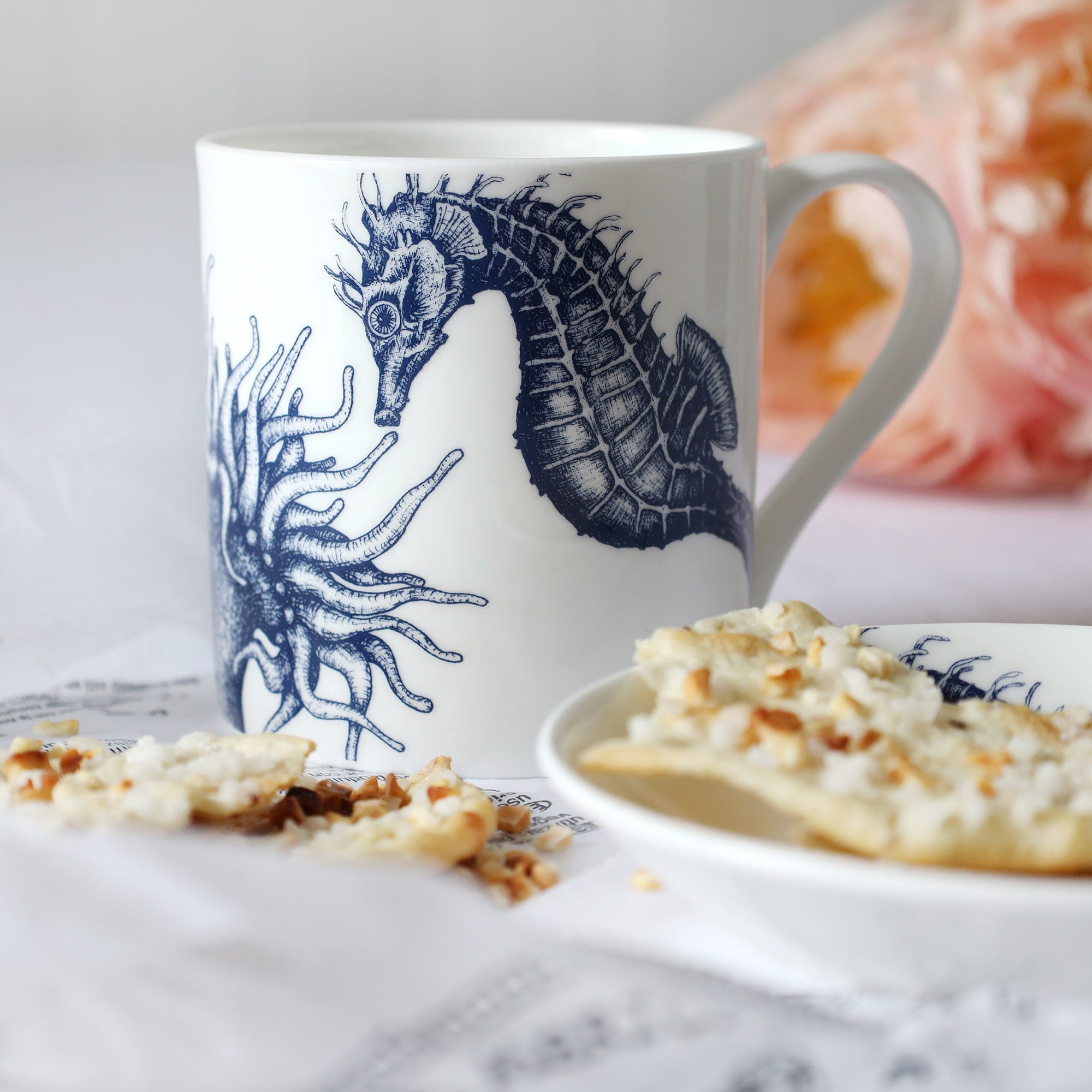 White mug with dark blue seahorse, anemone & scallop shell illustration sitting on a table with a plate & biscuits and flowers in the background.