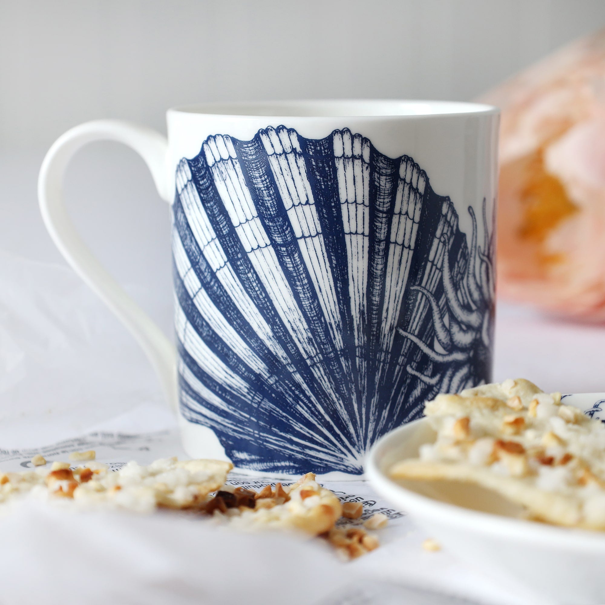White mug with dark blue scallop shell, anemone & seahorse illustration sitting on a table with a plate & biscuits and flowers in the background.