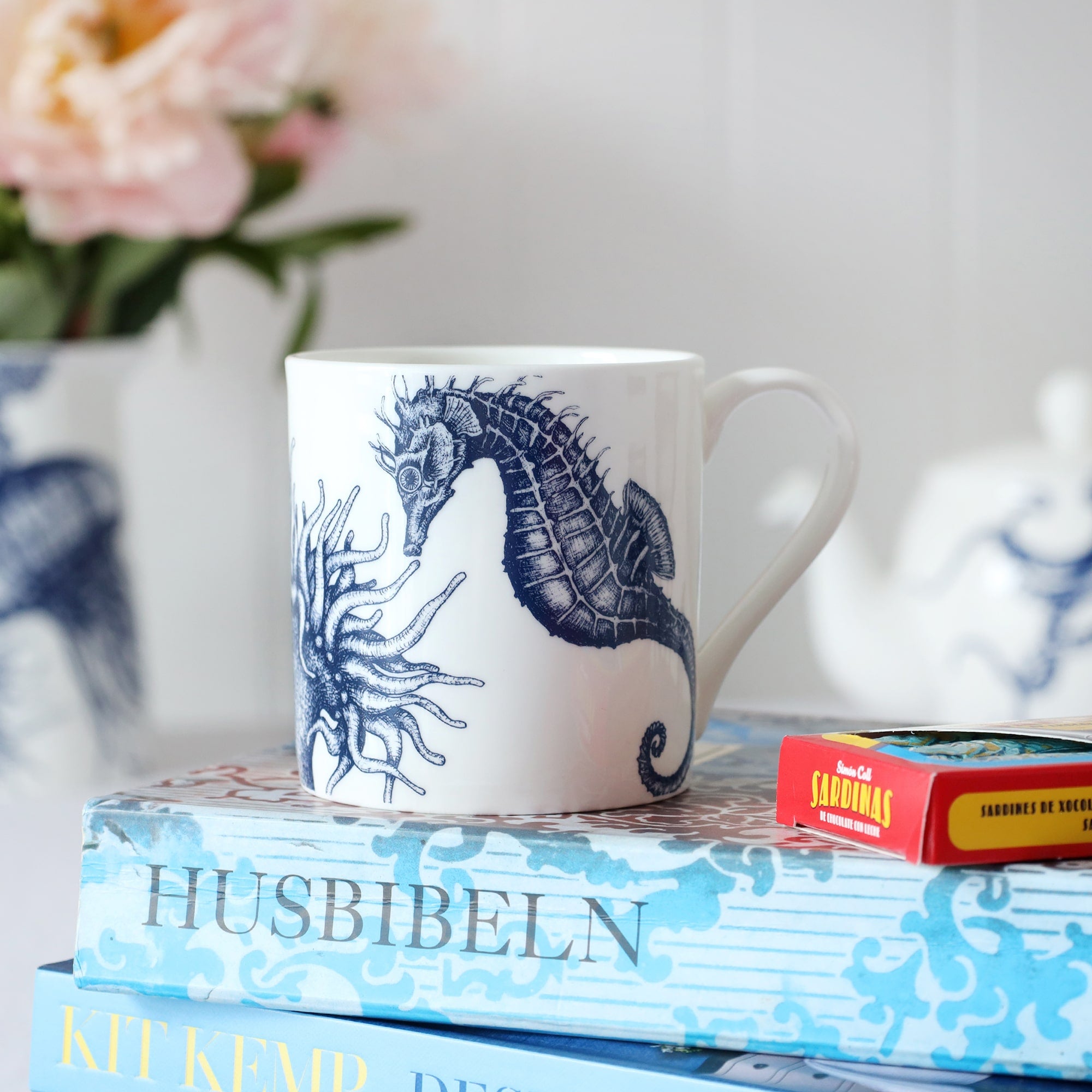 Close up of a white bone china mug with a blue illustrated seahorse & anemone on it, sitting on a table with biscuits and sardine chocolates and a pile of out of focus flowers and teapot in the background. 