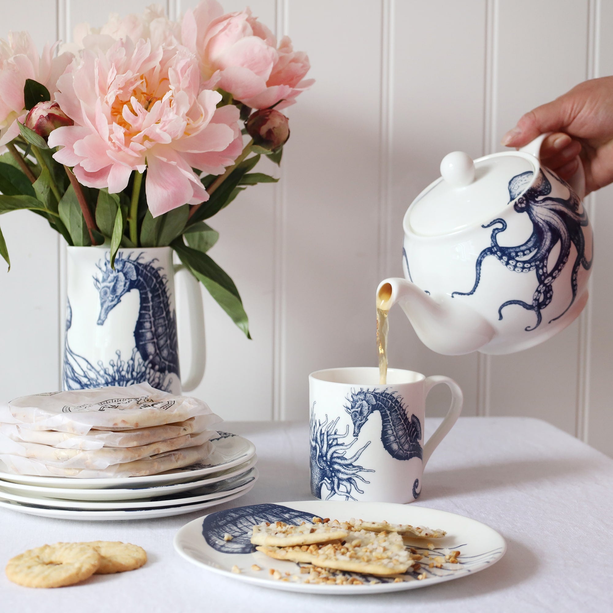 A blue & white informal; table setting set against a white shiplap wall and linen tablecloth. An octopus design adorns a teapot which is pouring tea into a white bone china mug decorated with a blue illustrated seahorse, anemone & scallop shell mug.
