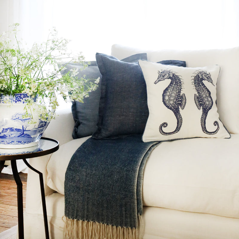 row of two blue cushions and a blue & white illustrated seahorse cushion at the front, sitting on a navy throw on a white sofa with bright sunlight window behind and a large willow pattern jug filled with cow parsley