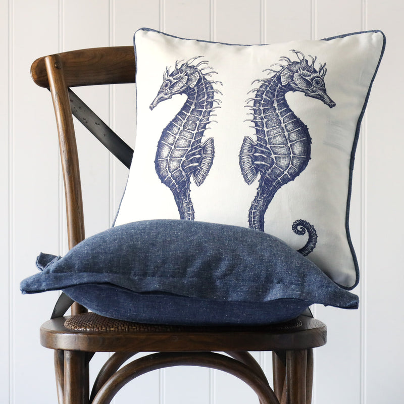 white cushion with navy illustrated two seahorse back to back on the front, placed on a linen cushion on a wooden chair