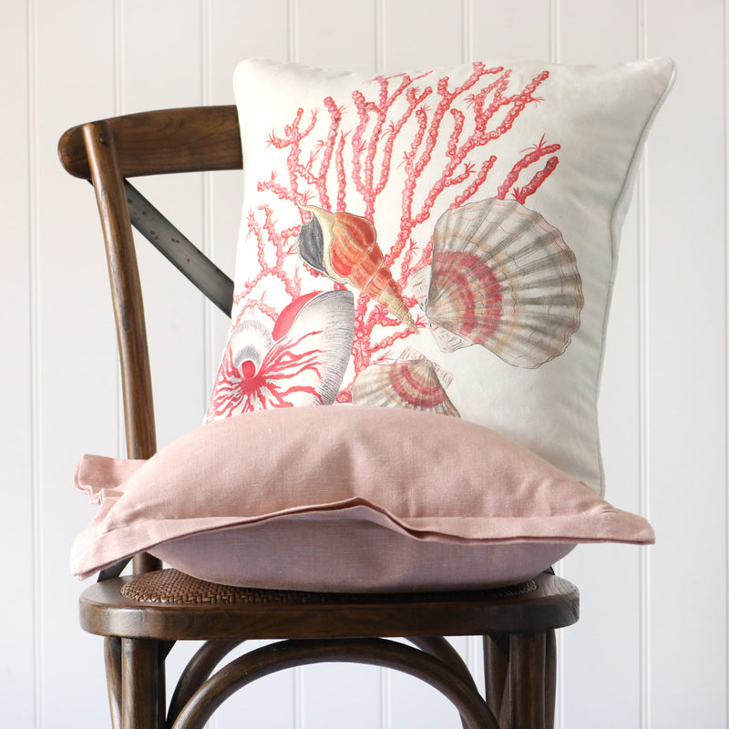 white cushion with illustrated nautilus shell, scallops and coral all in pink tones illustrated on the front, placed on a linen cushion on a wooden chair