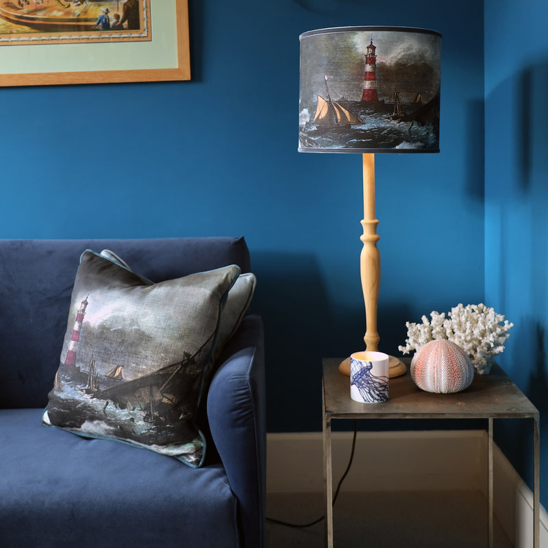 lampshade with shipwreck scene and a red and white lighthouse on wooden lamp base sitting on a metal table with coral, urchin and candle votive. There is a dark blue velvet sofa next to the table with a matching lighthouse cushion on it, and all set against a deep teal blue wall in the background.