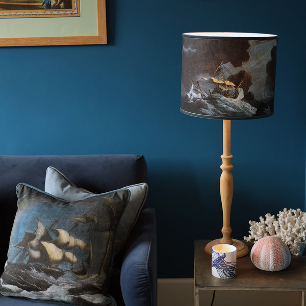 Lampshade with shipwreck scene on a wooden lamp base sitting on a table with sea urchin, coral & candle with a matching cushion on the sofa next to it