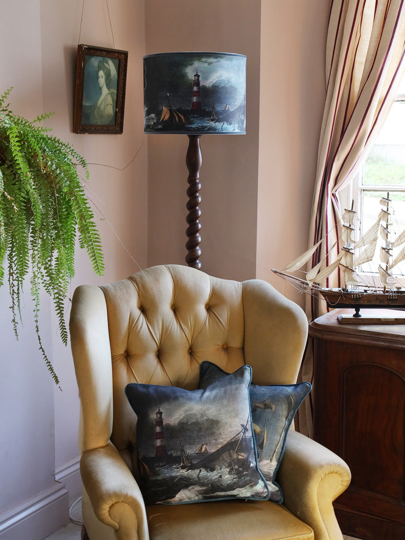 A shipwreck & lighthouse scene lampshade in blues & brown sitting on a petrol blue glass lamp base, set against a white shiplap wall a mustard throw over the chair and the corner of a matching cushion.