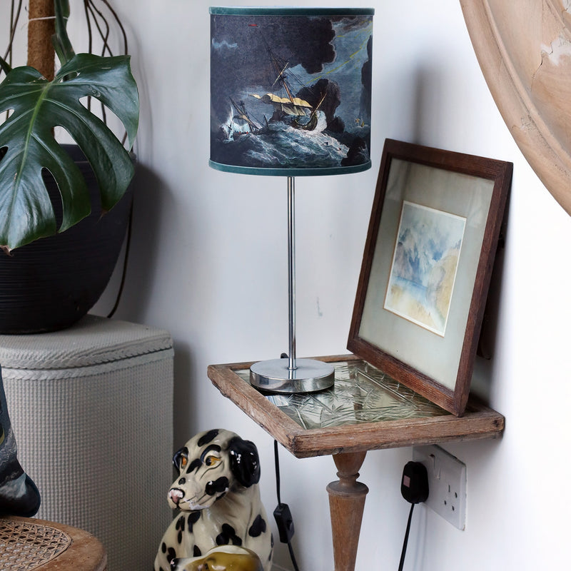 Small shipwreck scene lampshade and matching cushion set within a the corner of white room with an eclectic mix of various vintage objects and a large cheese plant.