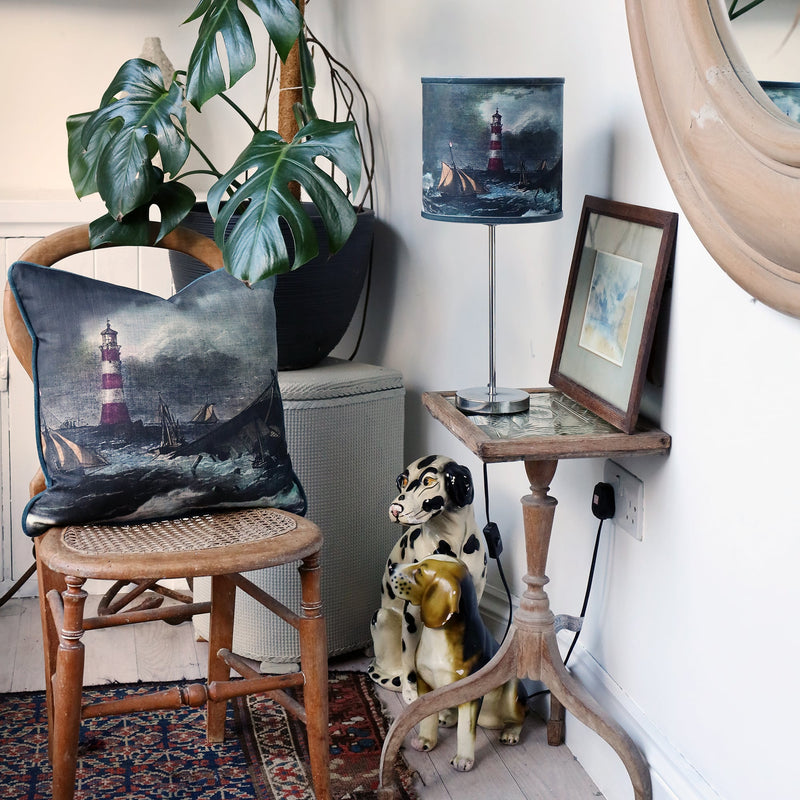 Small shipwreck scene lampshade and matching cushion set within a the corner of white room with an eclectic mix of various vintage objects and a large cheese plant.