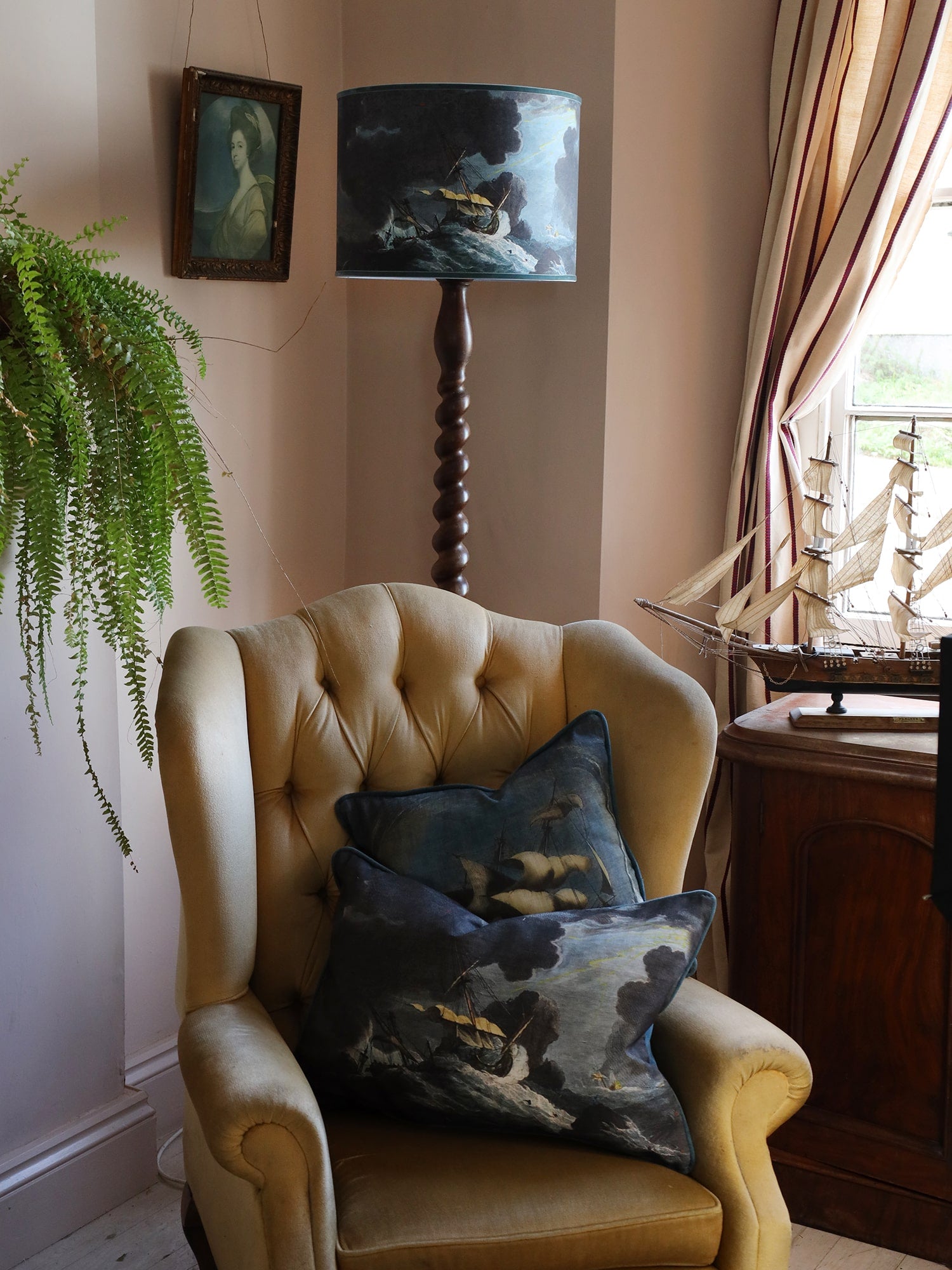 A shipwreck scene lampshade in dark blues and brown on a dark wooden twisted standard lamp base, placed behind a mustard velvet buttoned high back chair with an old portrait of a lady to the side and a model of a galleon on the table, all set against pink plaster walls. 