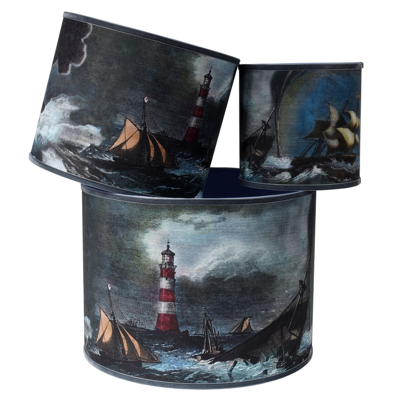 A stack of 3 different sized lampshades on white background, with nautical seascape images of a ships in distress in stormy seas and a red & white lighthouse in dark blues and browns and trimmed with a grey velvet ribbon.