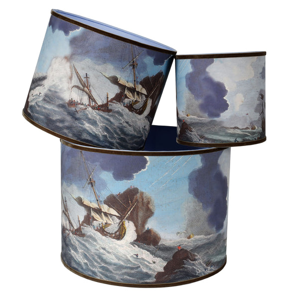 A stack of 3 different sized lampshades on white background, with nautical seascape images of a shipwreck in stormy seas in soft blues and trimmed with a mouse brown velvet ribbon.