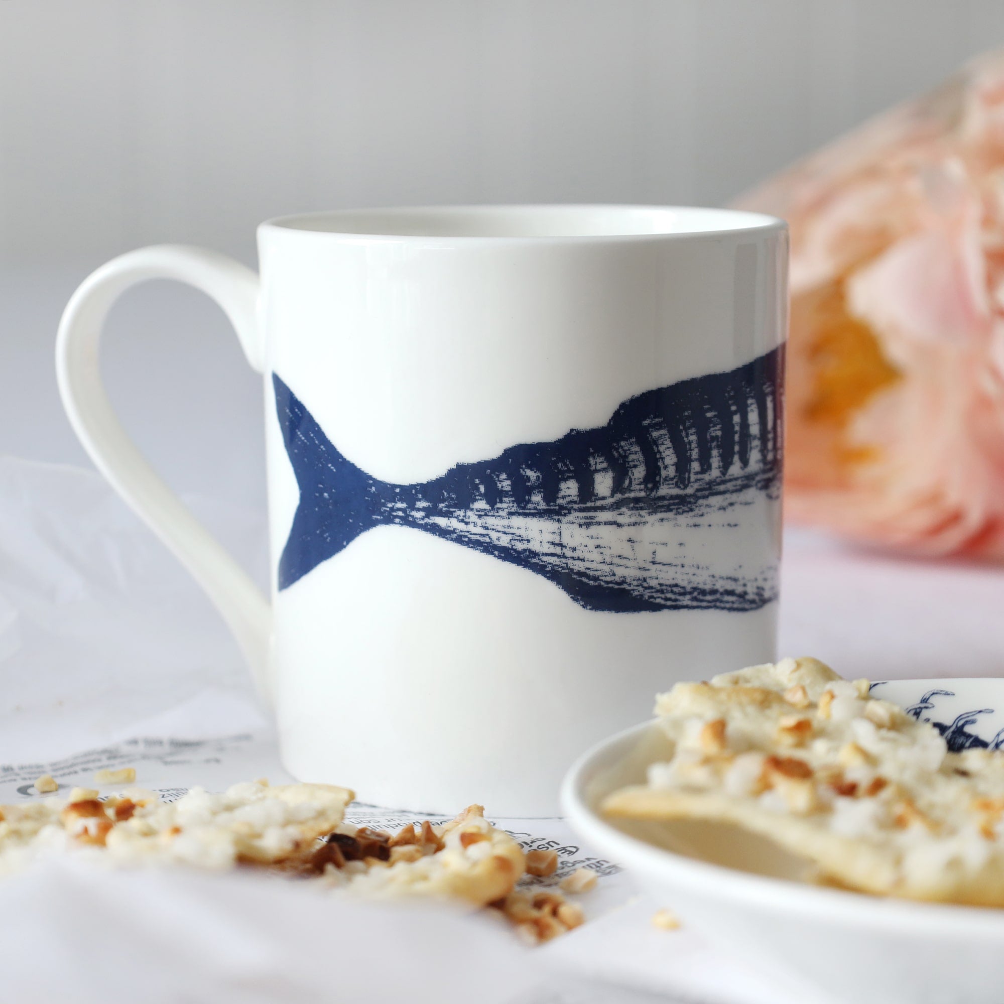 Blue & white bone china mackerel mug, with tail showing  on table with biscuits & flowers.
