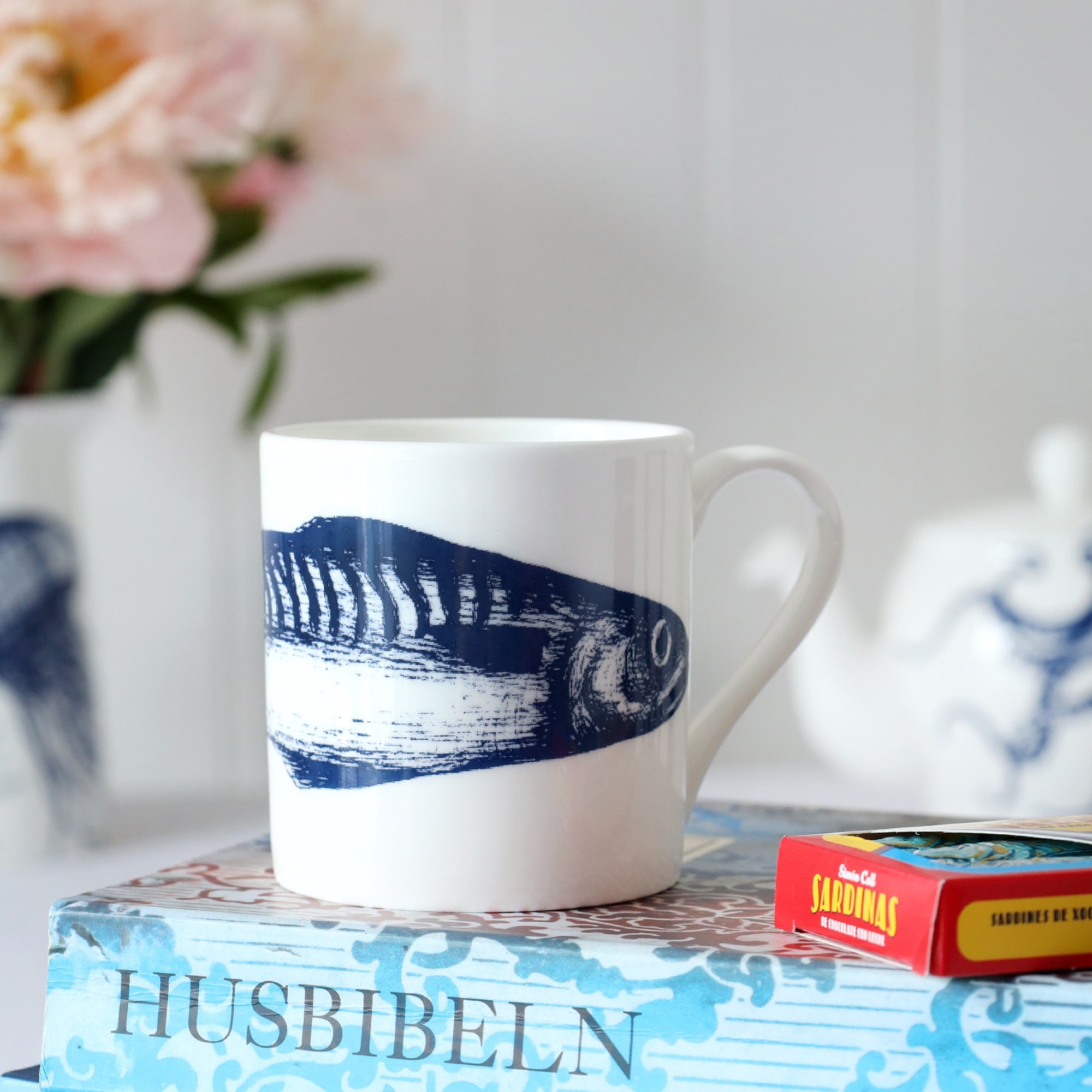 Blue & white bone china mug with a single mackerel around the middle sitting on top of a book with flowers in a jug in the background. 