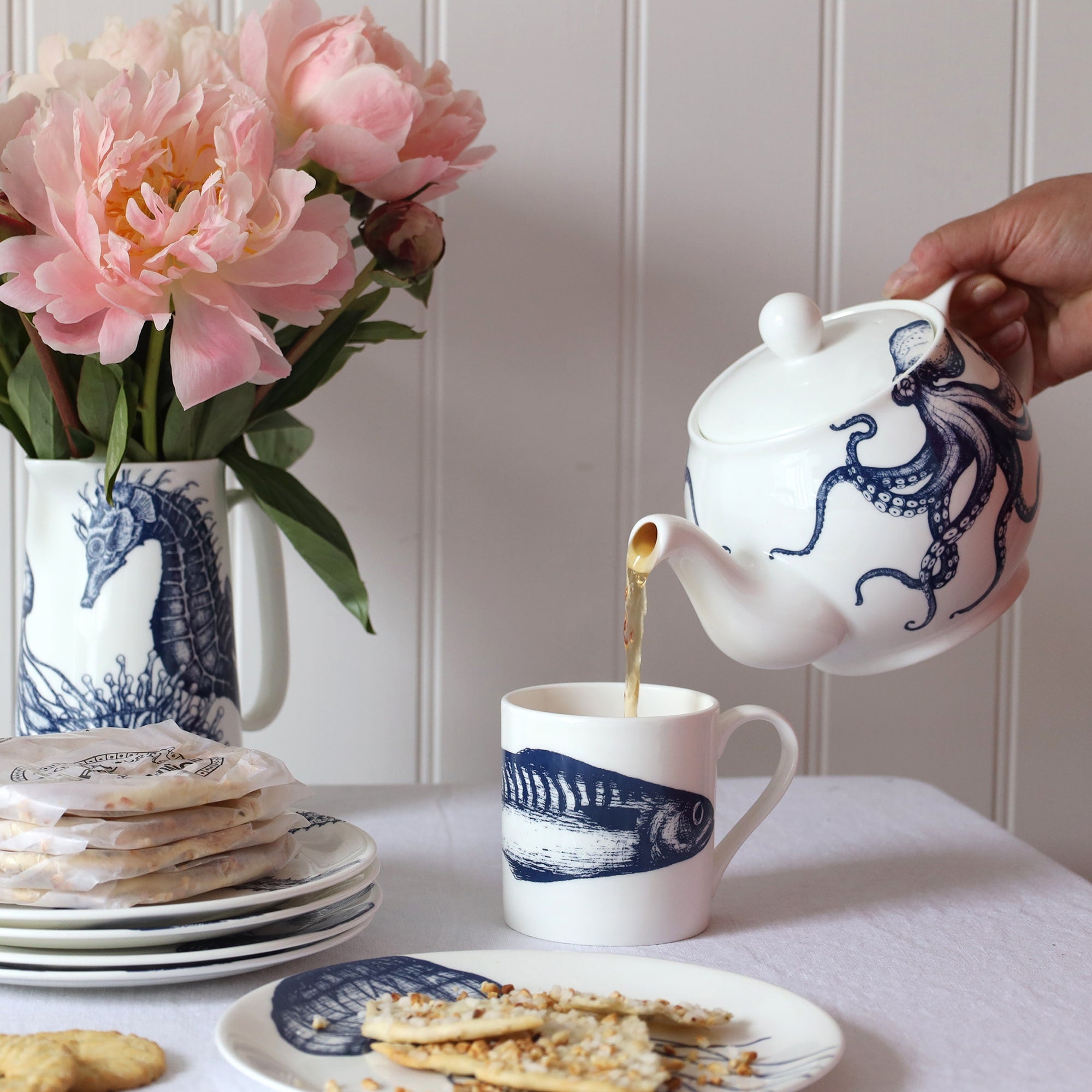 Table scene with blue & white coastal inspired plates & biscuits with a teapot pouring tea into a mug with single mackerel design around the middle. 
