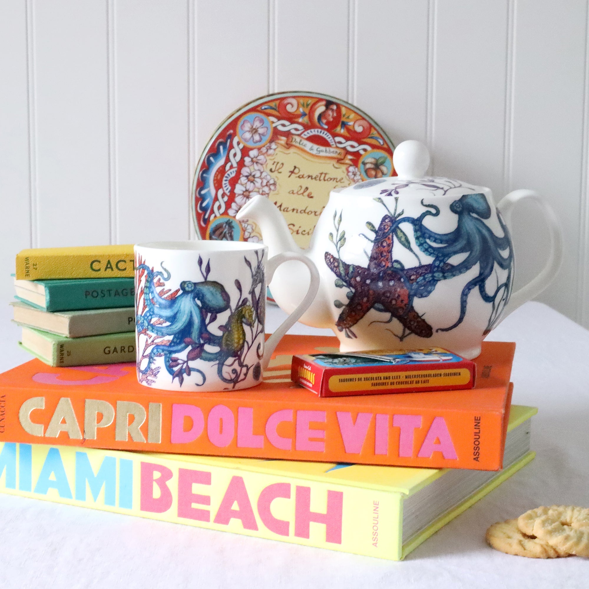  brightly coloured mug and teapot with reef design of many brightly coloured sea creatures. These are sitting on a table with brightly coloured books & panettone tin int he backgound.