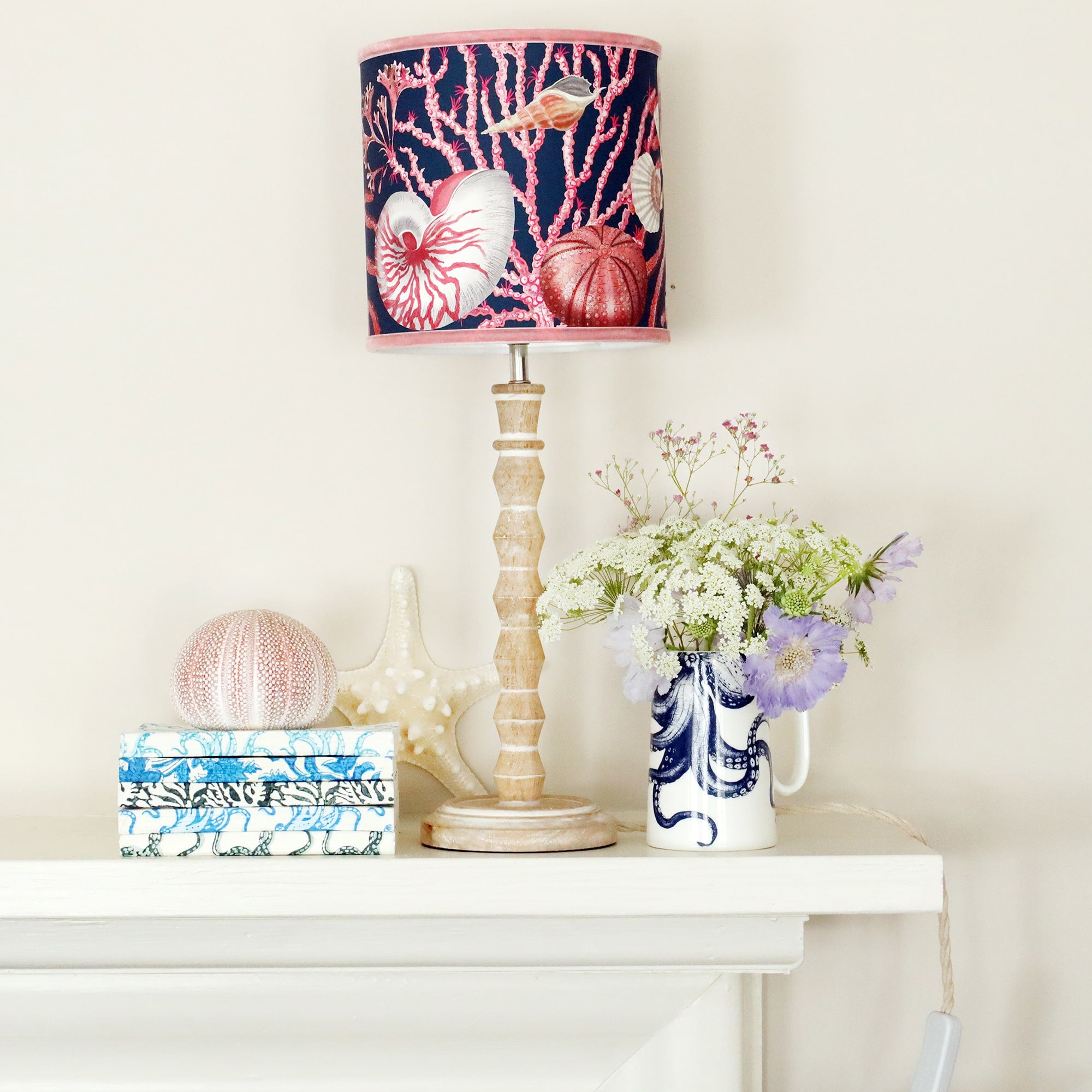 Whitewashed wooden small bobbin lamp base with a navy shell seeker lampshade, sitting on a mantelpiece with a jug of summer flowers a pile of block printed notebooks and beachcomber finds.