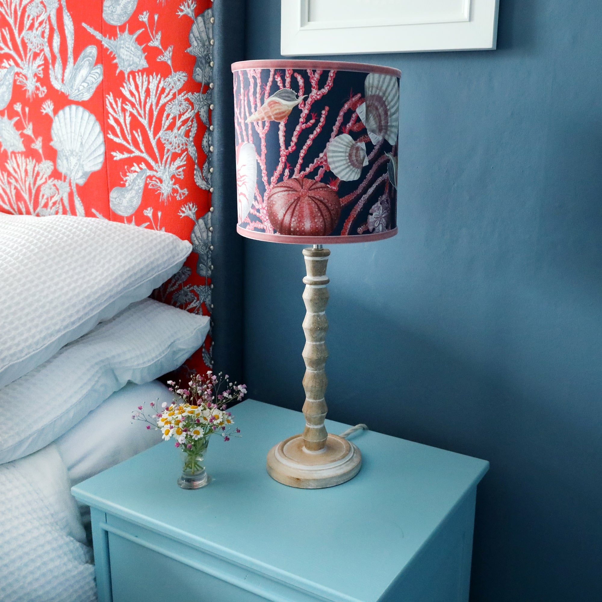 Whitewashed wooden small bobbin lamp base on set of light blue bedside drawers, dressed with a shell seeker navy lampshade and set beside a bed with a large shell fabric covered headboard.