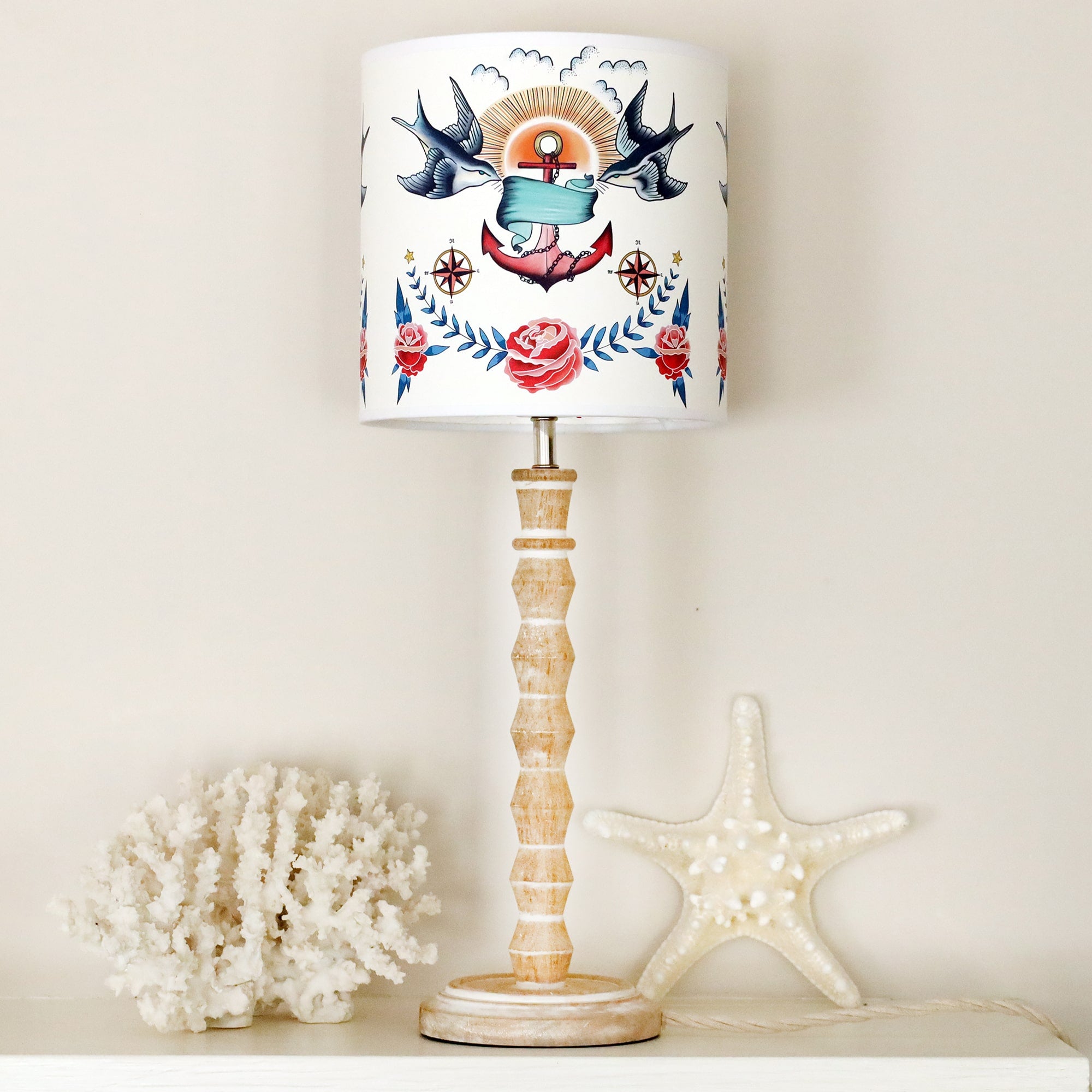 Whitewashed wooden small bobbin lamp base with tattoo inspired lampshade sitting on a mantlepiece with coral & starfish next to it.