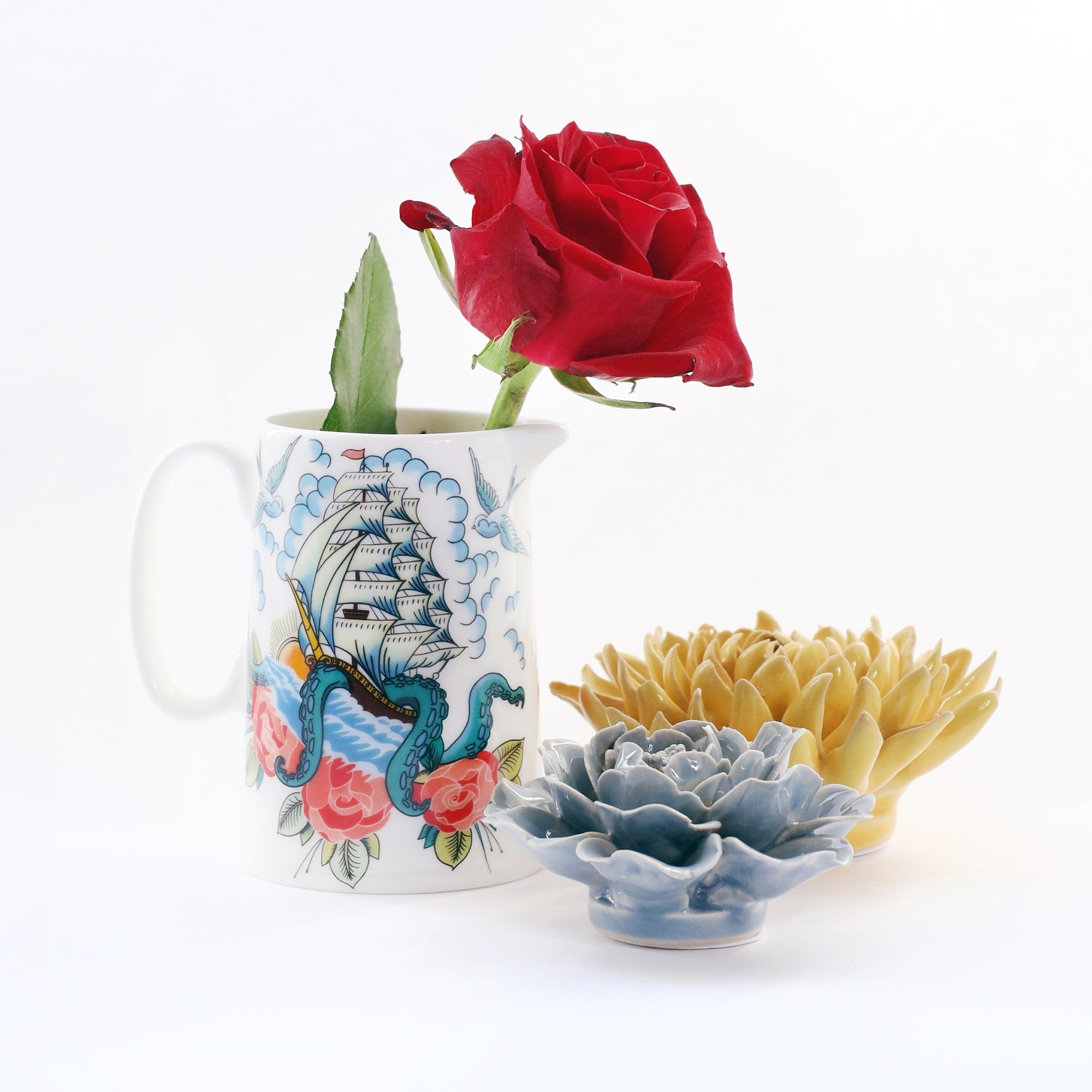 White jug with ship,kraken and roses design inspired by sailor's tattoos on a white background with 2 ceramic flowers nest to it and a red rose in the jug.
