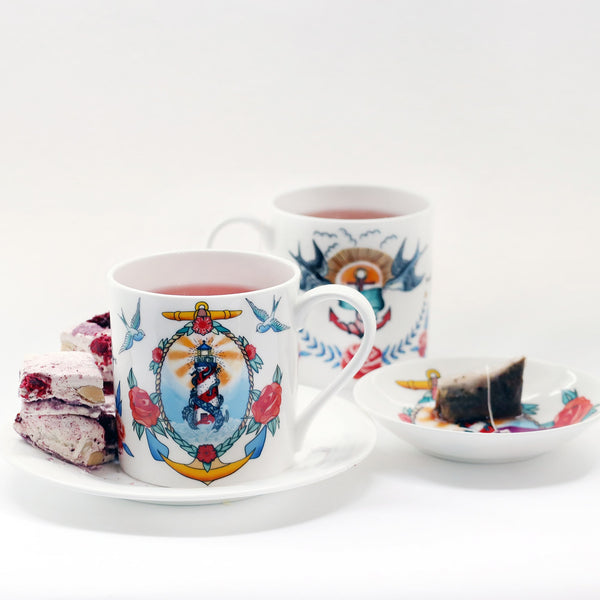 2 white mugs decorated in brightly coloured sailors tattoo inspired designs. The design in the foreground is a lighthouse and kraken surrounded with a rope, anchor and roses. This is sitting on a plain white plate with some pink nougat and there is a nibbles dish to the side with a tea bag in.