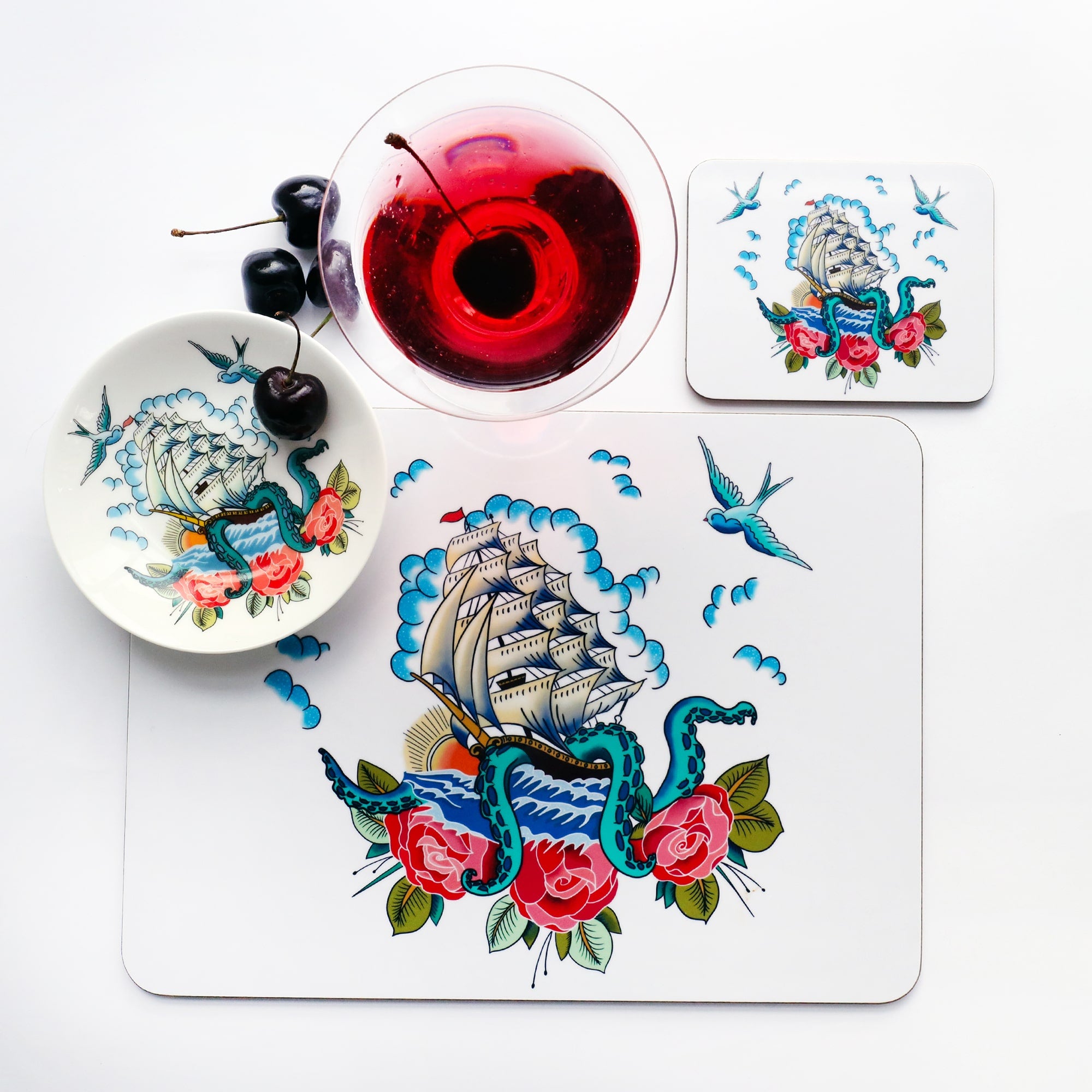 Place setting with placemat, coaster and nibbles dish all the the same ship, kraken and roses design that is inspired by a sailor's tattoos. The place setting is shot from above and there is a red cocktail and cherries just above the placemat.