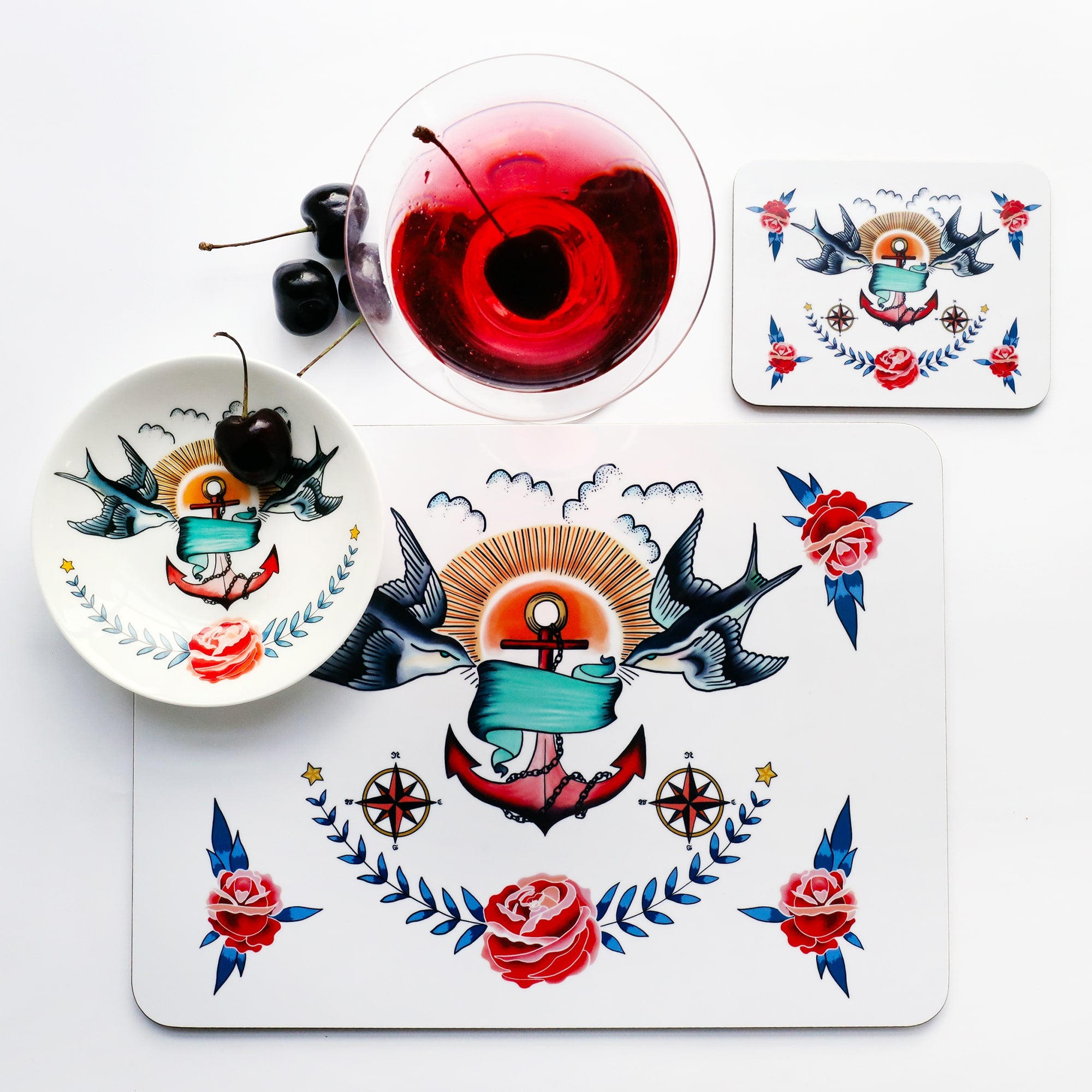 Place setting of placemat, coaster and nibbles dish with the same tattoo inspired design of swallows, anchor and rose. There is a red cocktail with a cherry in it and 3 cherries to the side.