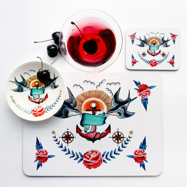 Place setting with placemat, coaster and nibbles dish all the the same swallows and anchor design that is inspired by a sailors tattoos. The place setting is shot from above and there is a red cocktail and cherries just above the placemat.