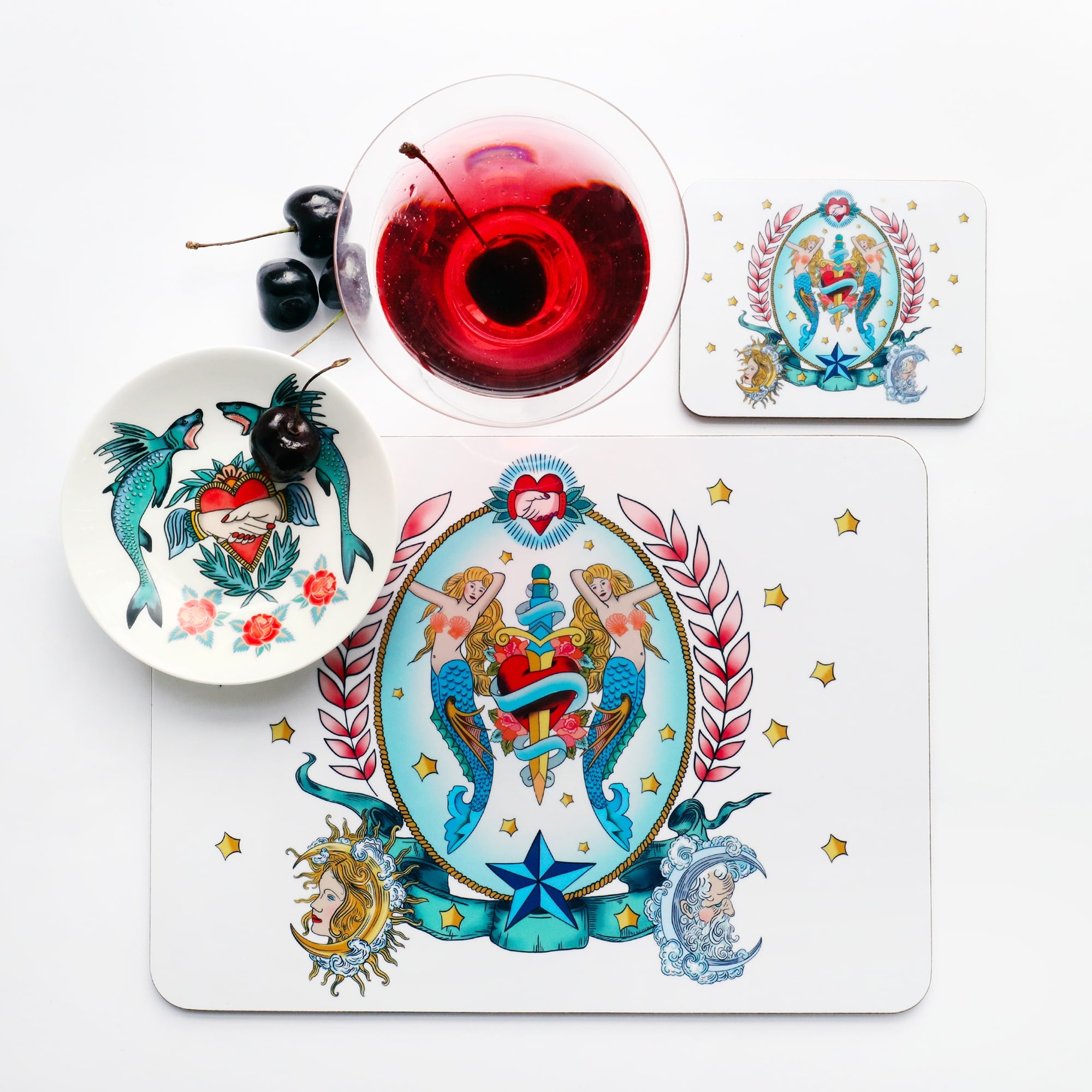 Place setting with placemat, coaster and nibbles dish all the the same mermaid, heart and dagger design that is inspired by a sailors tattoos. The place setting is shot from above and there is a red cocktail and cherries just above the placemat.