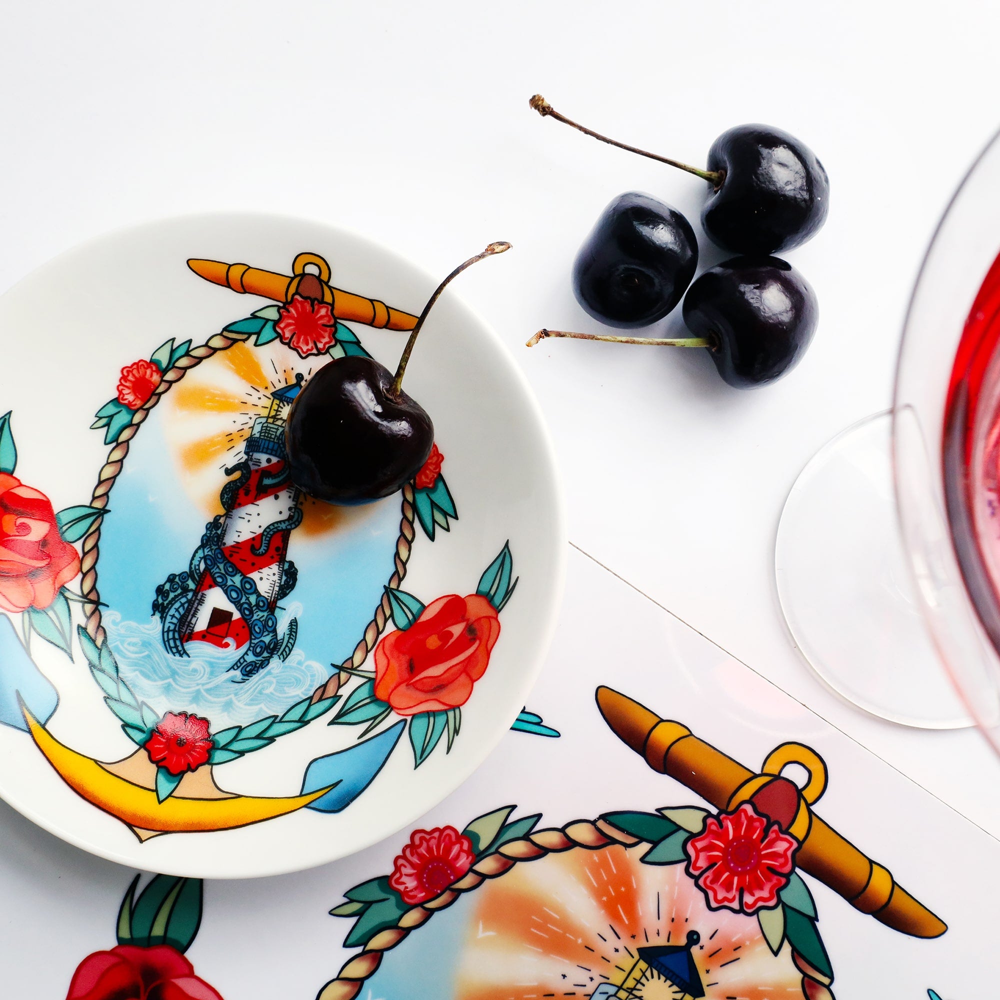 Nibbles dish with an illustration of a lighthouse  with kraken tentacles coming out, surrounded by a rope,anchor and roses. There is a cherry in the dish and 3 others next to it, you can also see a small amount of a placemat with the same design and the top of a cocktail glass just barely visible.