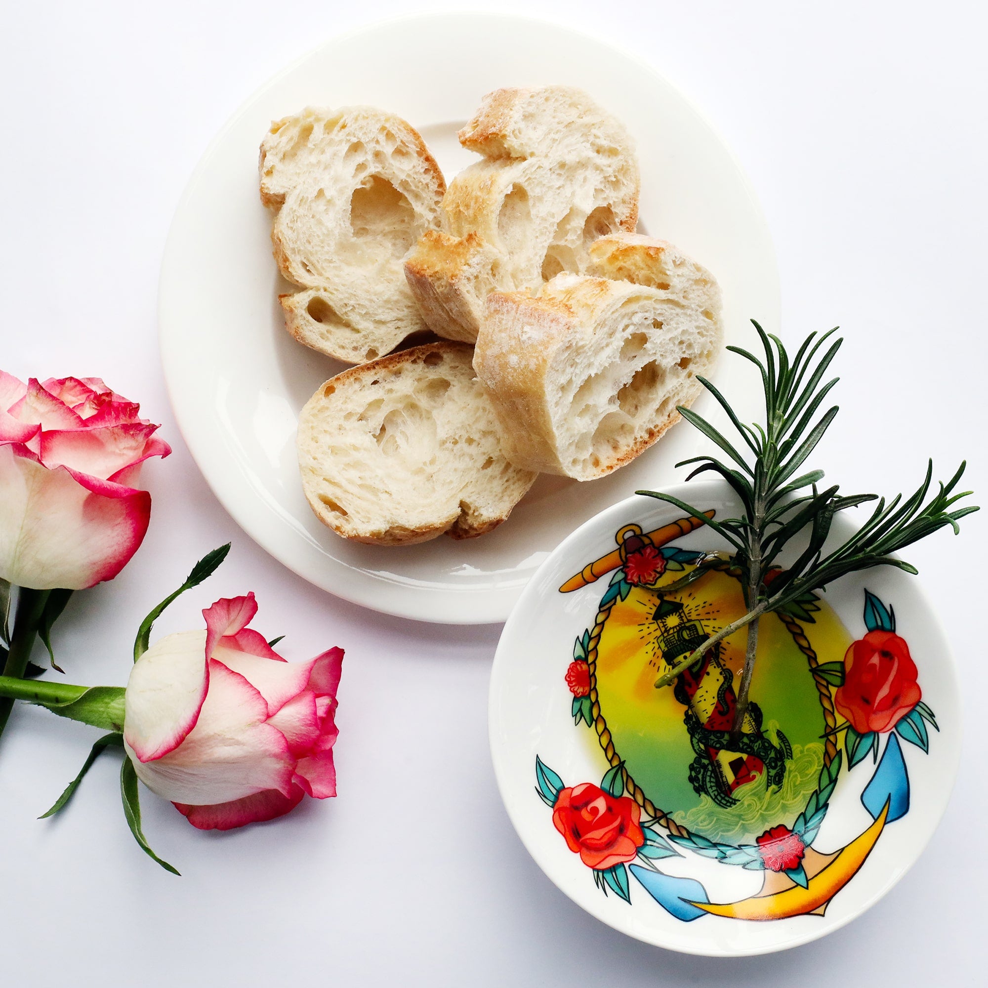 Nibbles dish with lighthouse, kraken, rose and anchor design in a tattoo style. the dish has olive oil and rosemary in it and there is a plate of bread cut to the side as well as 2 pink and white roses.