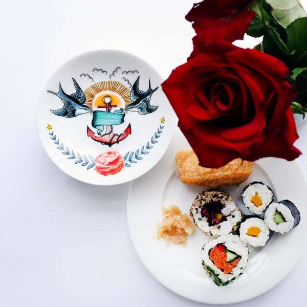 Nibbles dish decorated with tattoo inspired design of swallows, anchor and rose. There is a plate of sushi next to it and red roses.