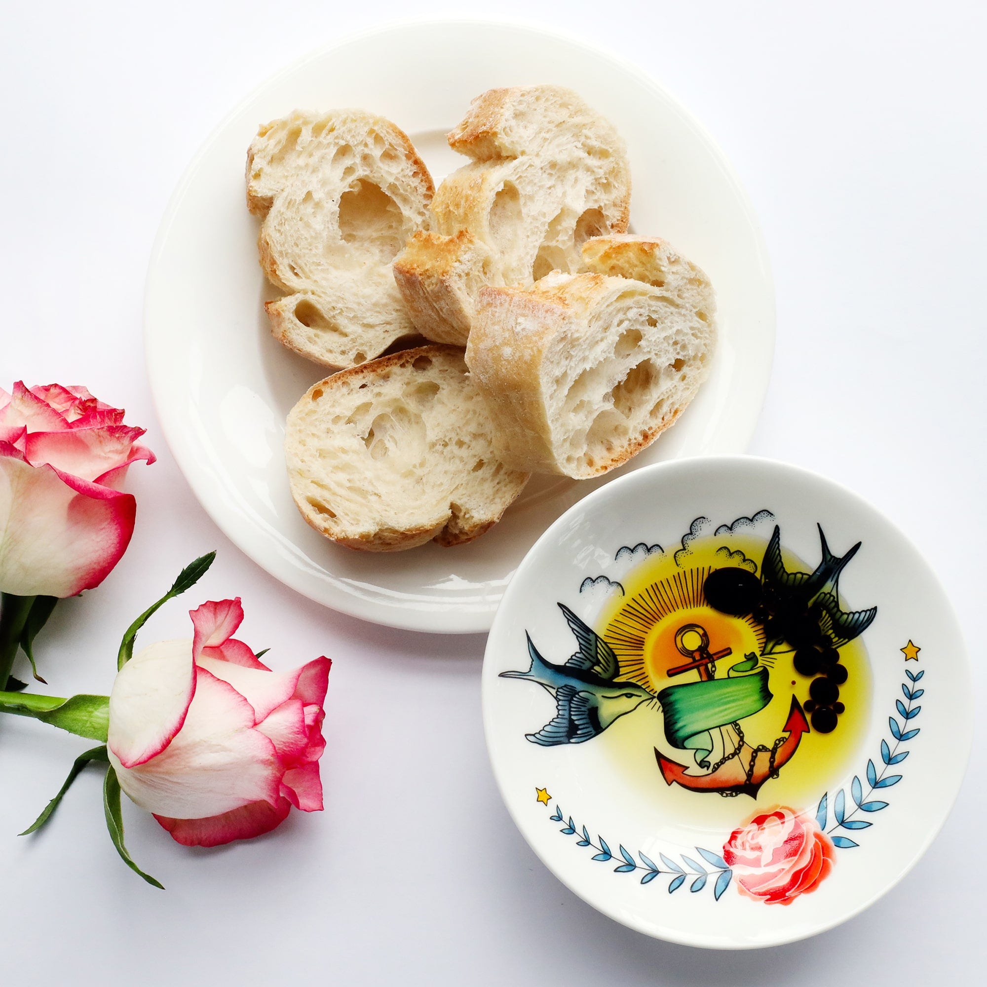 Nibbles dish with tattoo inspired design of swallows, anchor and rose, filled with olive oil and balsamic vinegar. There is a plate of bread to the side and 2 pink and white roses.