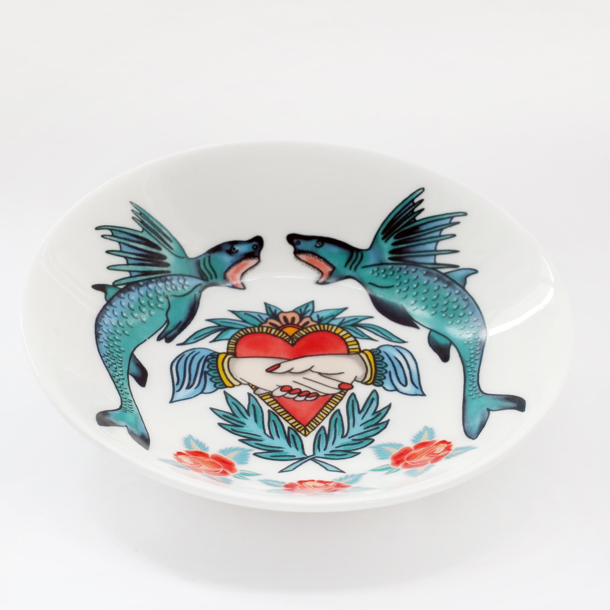 Product shot of a white bone china nibbles dish decorated with sharks, heart and hands and roses in bright tattoo colours.