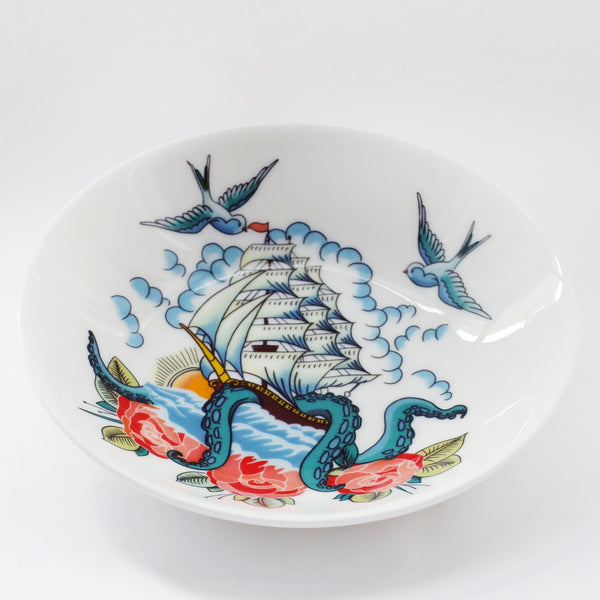 Product shot of a white nibbles dish decorated with a tattoo inspired ship & kraken design. There are roses at the bottom and doves flying at the top.