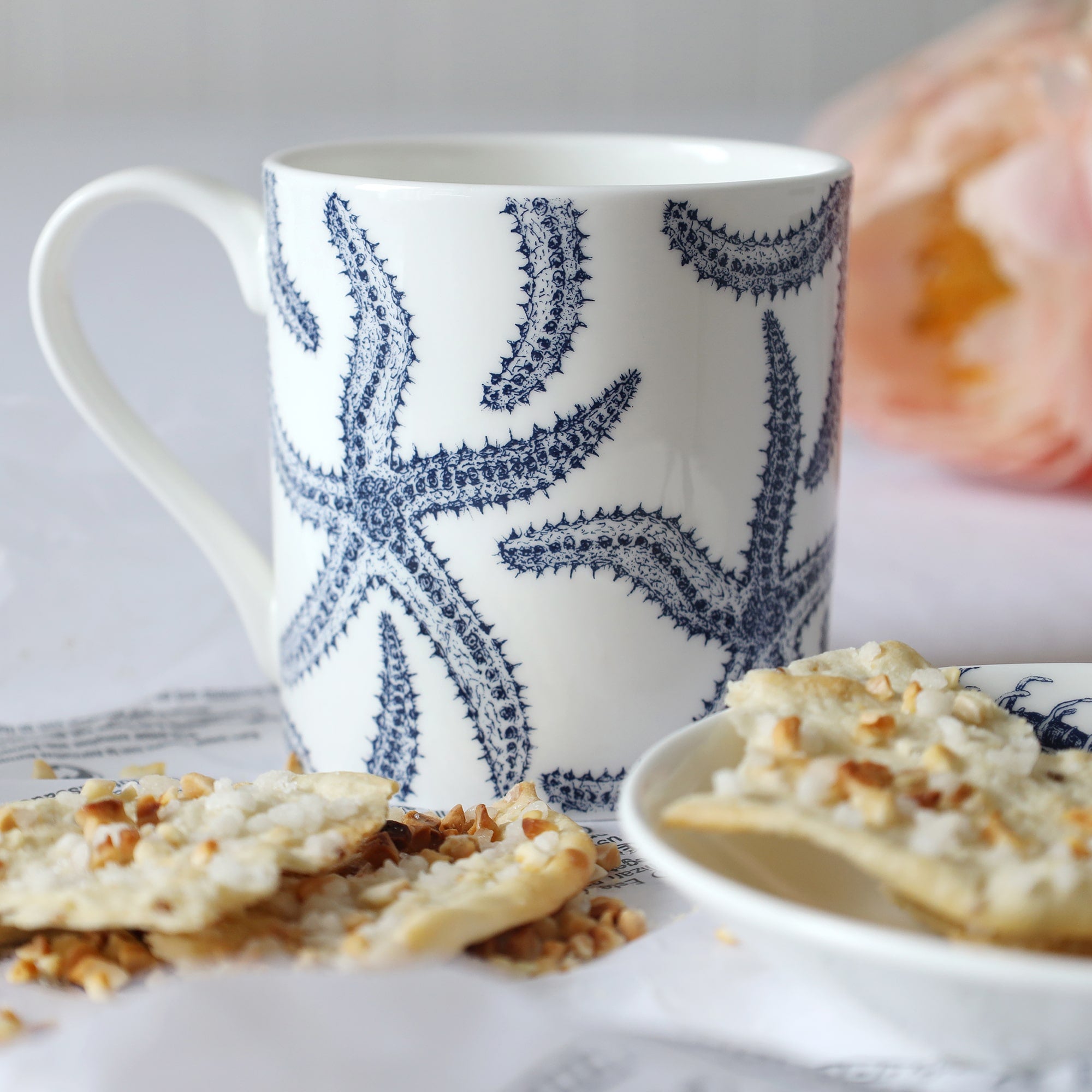 White mug with dark blue multiple starfish illustration sitting on a table with a plate & biscuits and flowers in the background.
