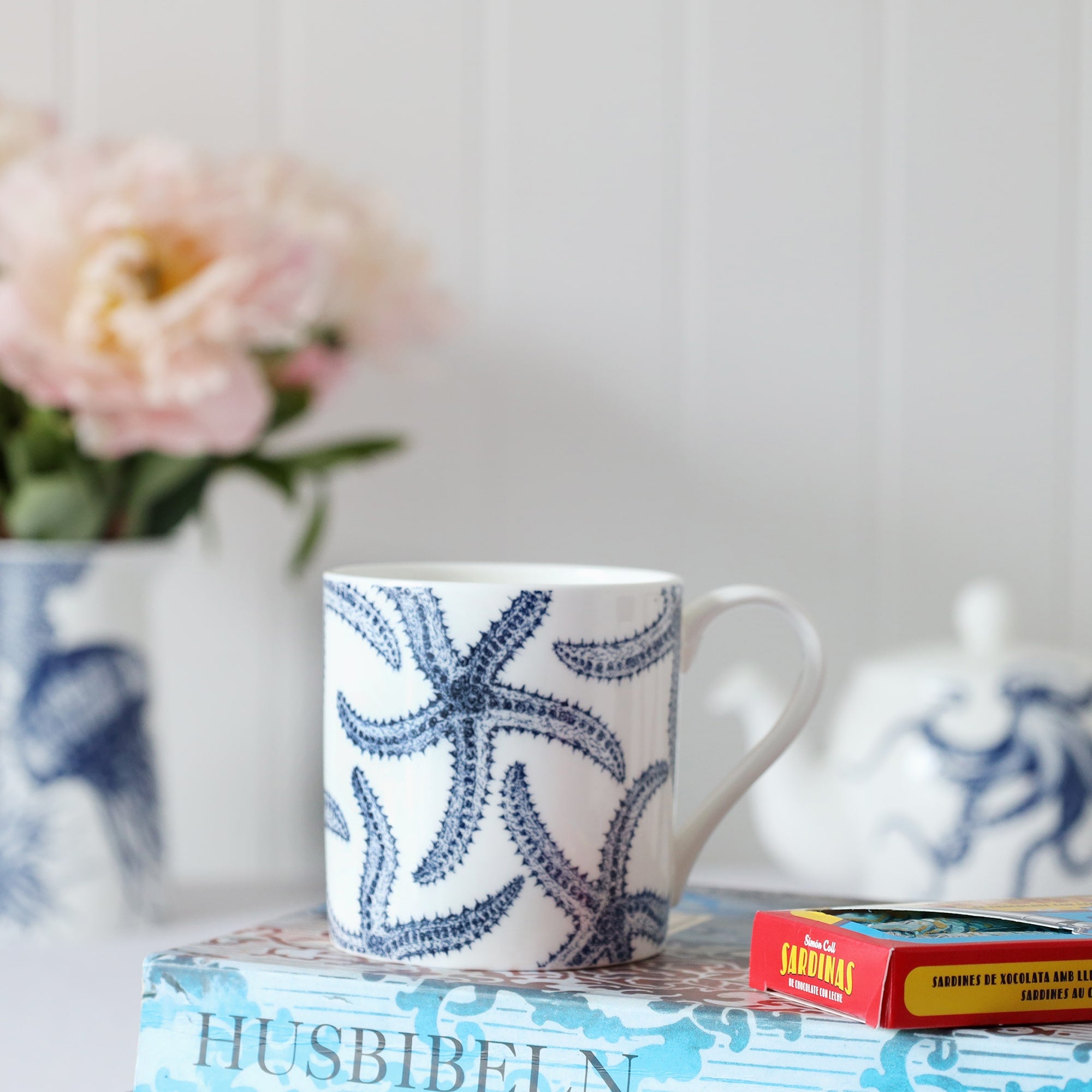 A blue & white informal table setting set against a white shiplap wall and linen tablecloth. An octopus design adorns a teapot which is pouring tea into a white bone china mug decorated with a blue illustrated multi starfish design..