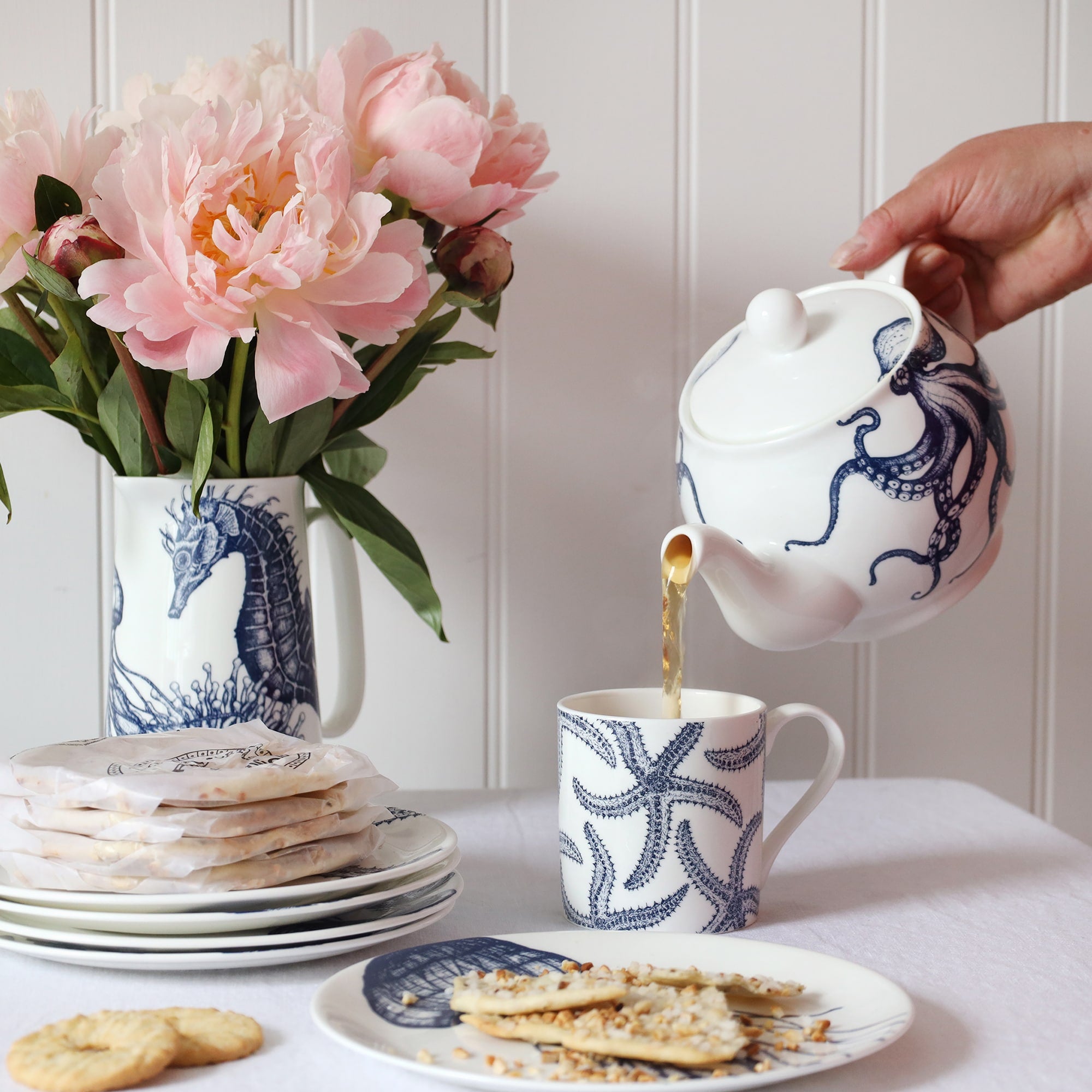 A blue & white informal table setting set against a white shiplap wall and linen tablecloth. An octopus design adorns a teapot which is pouring tea into a white bone china mug decorated with a blue illustrated multi starfish design..