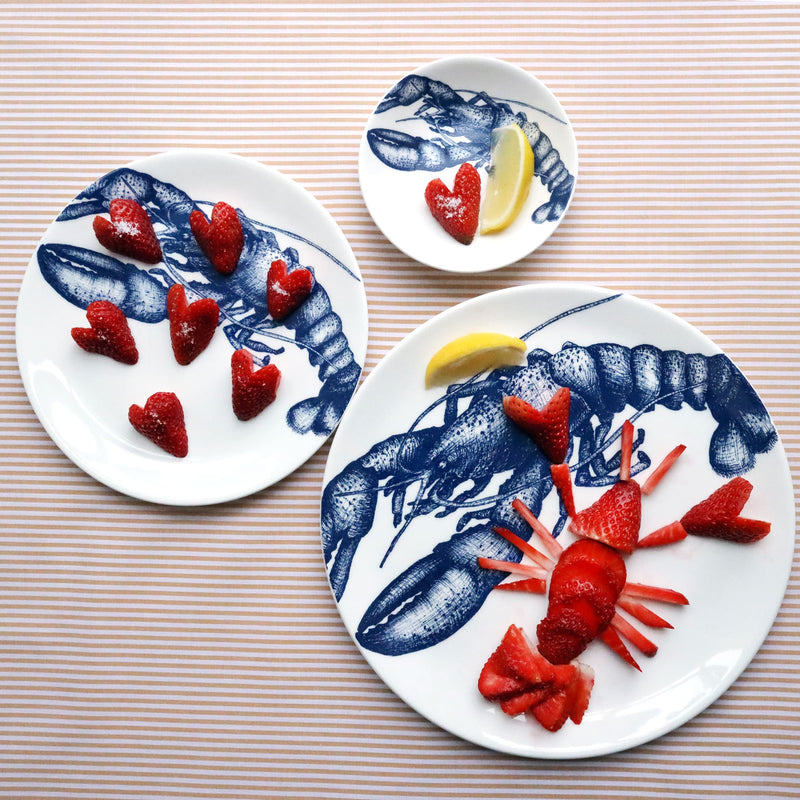 Three different sized white bone china plates with an illustrated navy blue lobster on them. The largest plate has a lobster made from strawberries on it and the other 2 have hearts made from strawberries and a slice of lemon on them. All three plates are shot from above on a striped yellow gold cloth.