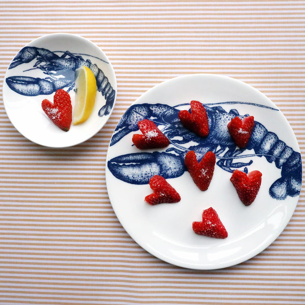 Two different sized white bone china plates with an illustrated navy blue lobster on them. The largest plate has hearts made from strawberries on it and the other 2 has a heart made from strawberries and a slice of lemon on it. Both plates are shot from above on a striped yellow gold cloth.