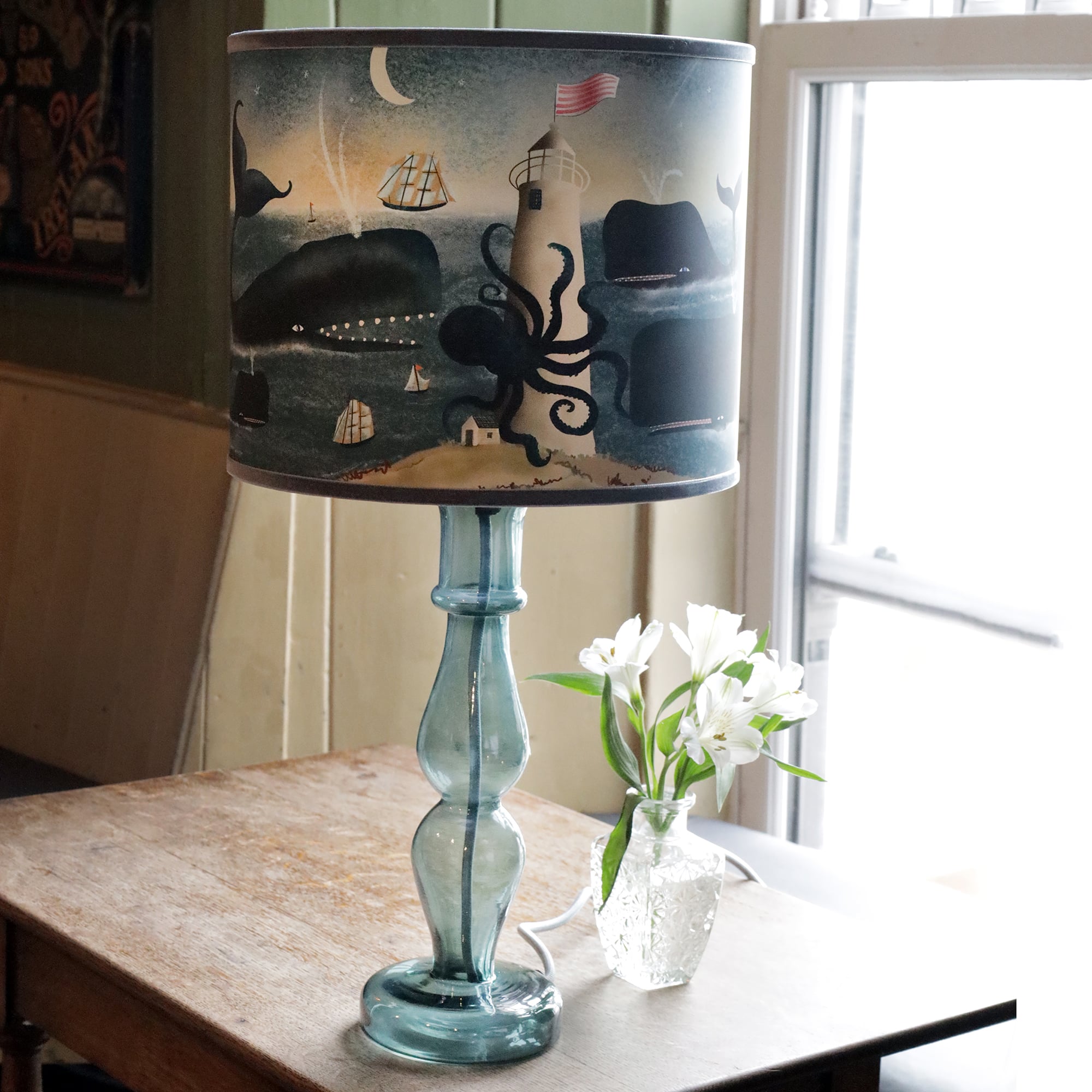 A blue glass lamp with a whimsical nautical-themed lampshade sits on a wooden table next to a vase of white flowers. The lampshade depicts an ocean scene featuring a lighthouse wrapped by a giant octopus, ships with billowing sails, whales, and a crescent moon in the sky. The soft natural light coming through a nearby window adds a calm and cozy atmosphere to the rustic room, complementing the sea-inspired decor.