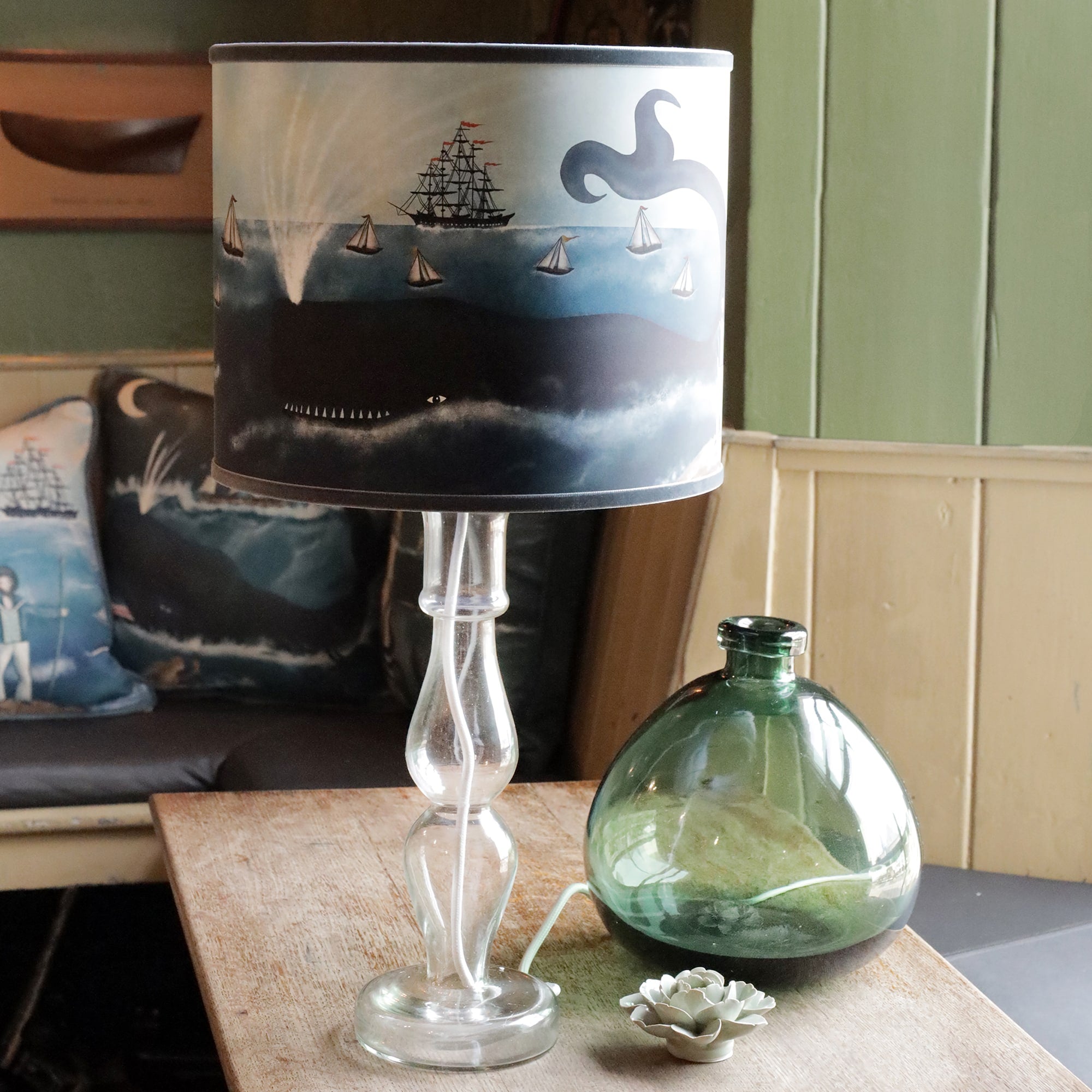 A glass lamp with a unique design sits on a wooden table. The lampshade features a maritime illustration showing an old ship with red flags surrounded by smaller sailboats and a large whale surfacing beneath the water, its tail raised in the air. Next to the lamp is a green glass vase and a small ceramic succulent. In the background, a couch adorned with pillows displays similar nautical imagery, reinforcing the oceanic theme in the cozy, rustic setting.