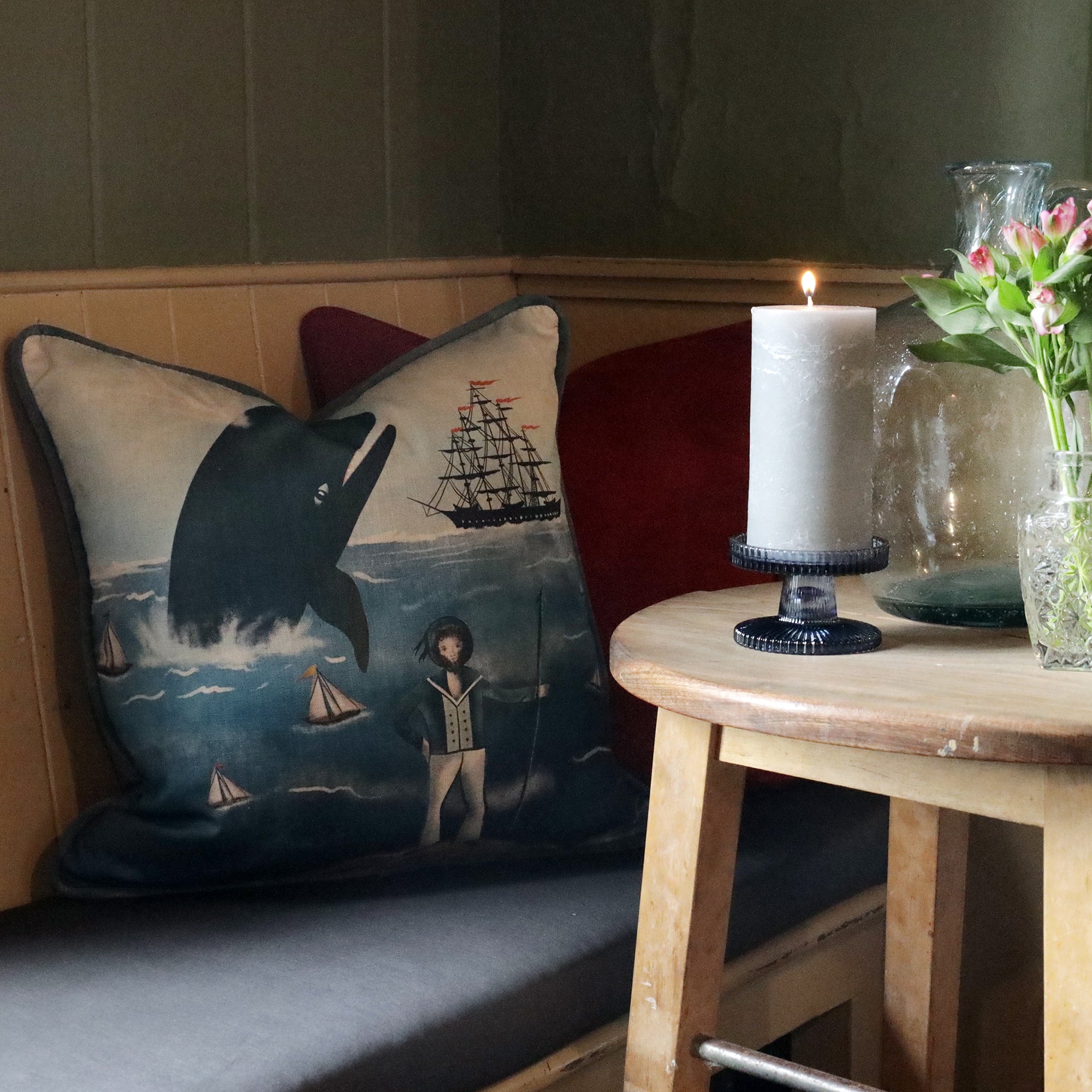 A cozy nook featuring a cushion with a nautical-themed design of a whale, sailboats, and a captain standing on the shore. The cushion rests on a bench alongside a red pillow, adding a pop of color to the space. In the foreground, a wooden side table holds a lit candle on a black holder, a vase of pink flowers, and a large glass jug, contributing to the serene and warm atmosphere. The soft lighting and maritime decor create a peaceful, seaside-inspired ambiance perfect for relaxation.