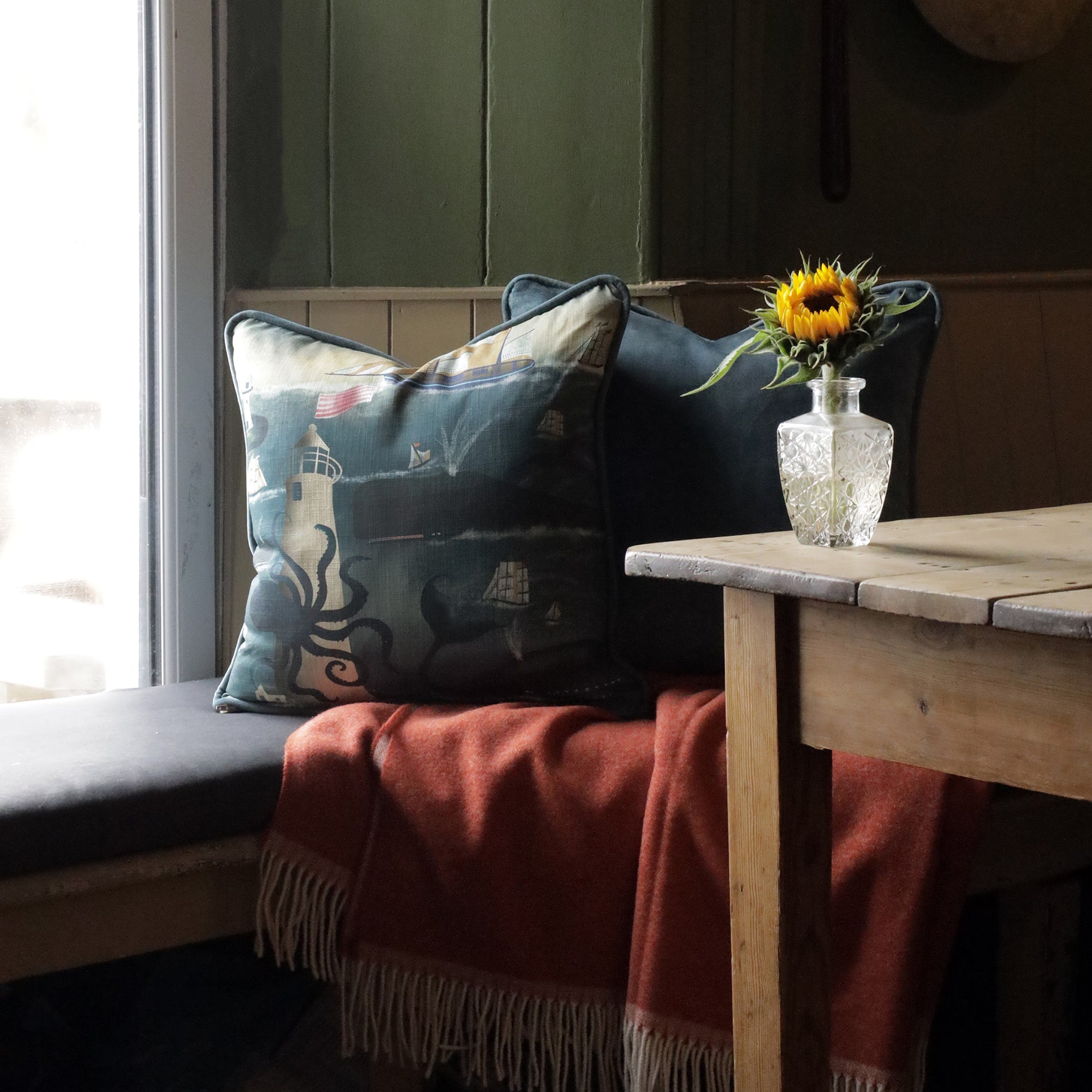 A cozy seating area featuring a nautical-themed cushion with an illustration of a lighthouse, octopus, whales, and sailboats on a deep blue ocean. The cushion is paired with a solid dark green pillow and draped over a soft orange throw blanket with fringe. A wooden table sits beside the bench, holding a small crystal vase with a bright sunflower. Soft natural light filters in through a nearby window, creating a warm, inviting atmosphere with a seaside charm.