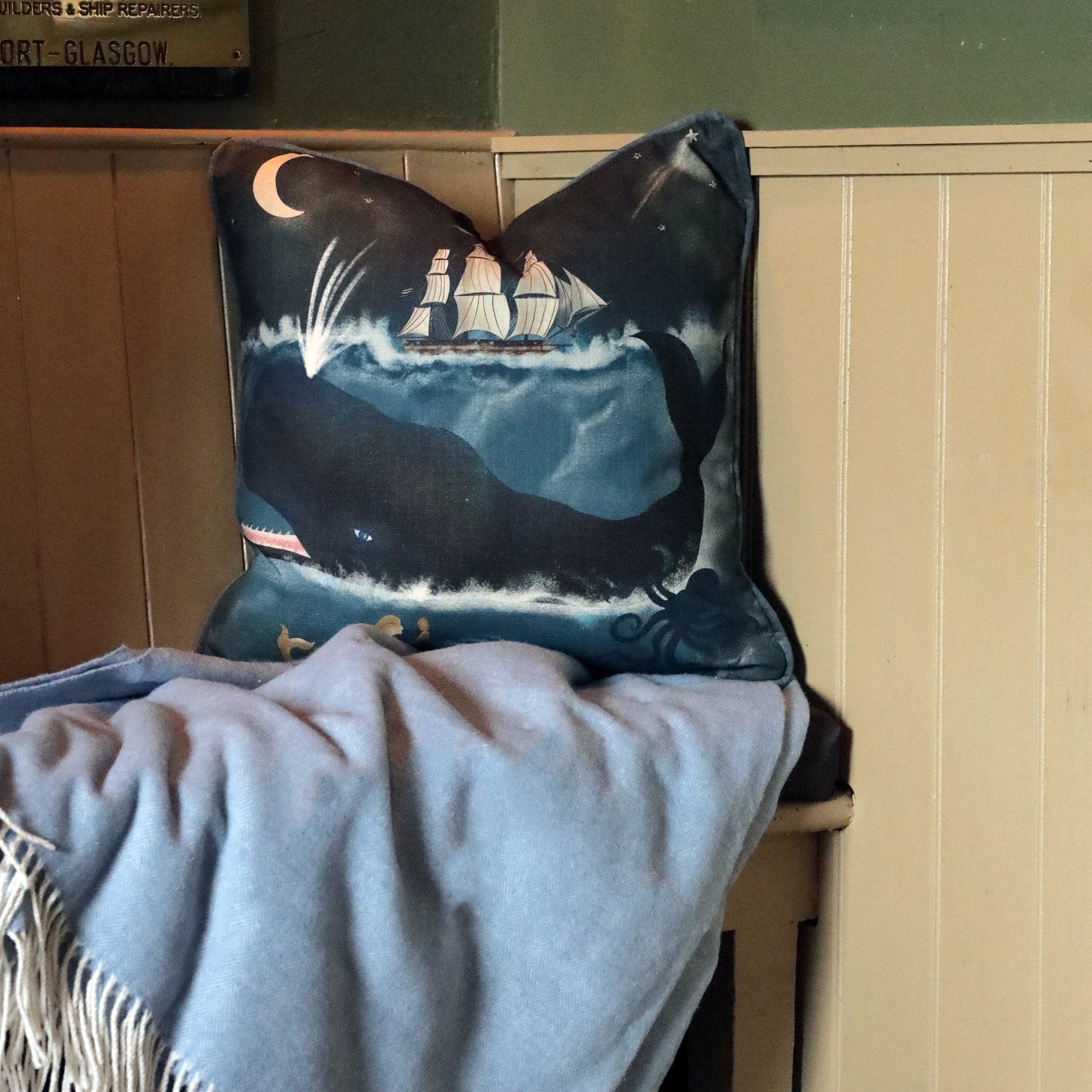 A cozy bench featuring a nautical-themed cushion with a design of a whale swimming beneath a tall ship under a crescent moon. The cushion is placed against a soft blue blanket, adding warmth and comfort to the space. The deep blue tones of the cushion and blanket complement the wooden paneling on the wall, creating a serene and inviting atmosphere. The maritime decor evokes a sense of coastal adventure, perfect for a relaxed, seaside-inspired setting.