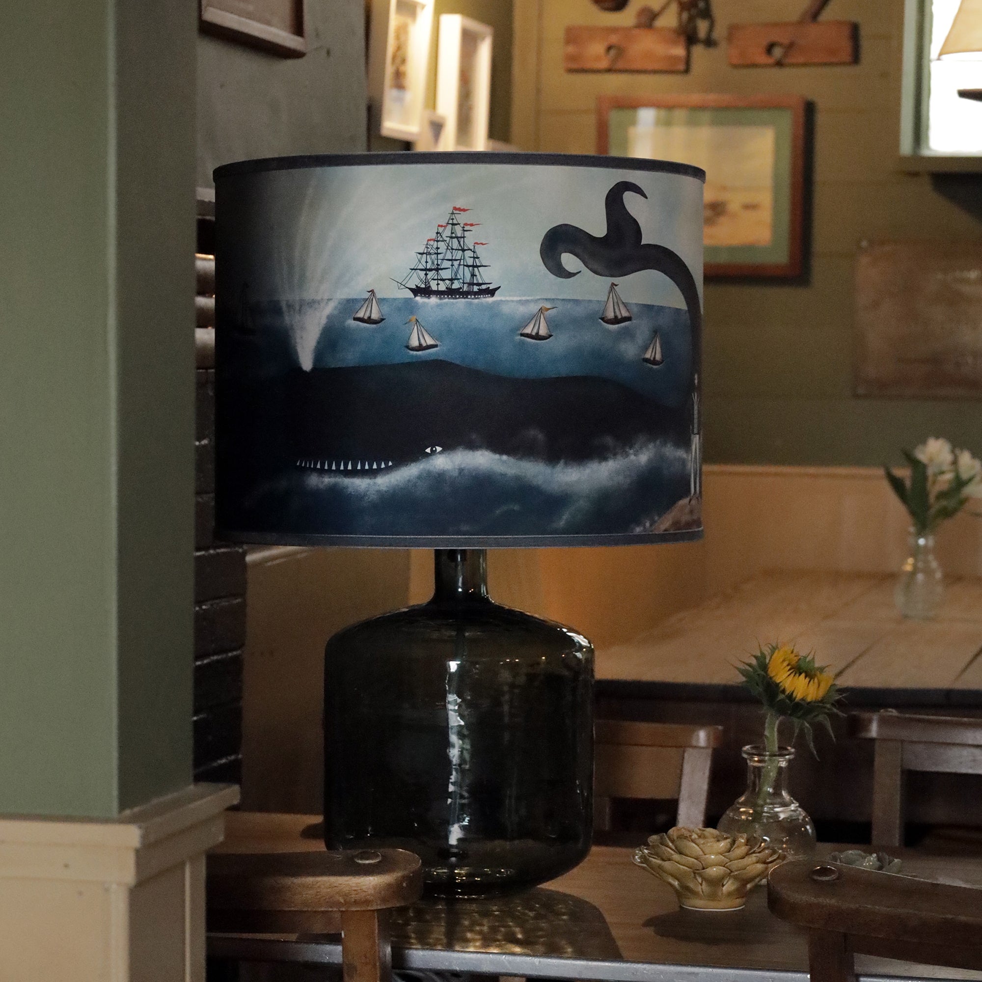 A large, dark glass lamp with a nautical-themed lampshade sits on a wooden table. The lampshade illustrates a scene of a sailing ship with red flags and smaller sailboats navigating the ocean, while a large whale surfaces below, spouting water. A small sunflower in a glass vase and a decorative artichoke-shaped dish are placed beside the lamp. In the background, framed maritime art and wooden pulleys hang on the walls, creating a cozy, sea-inspired atmosphere in the room.