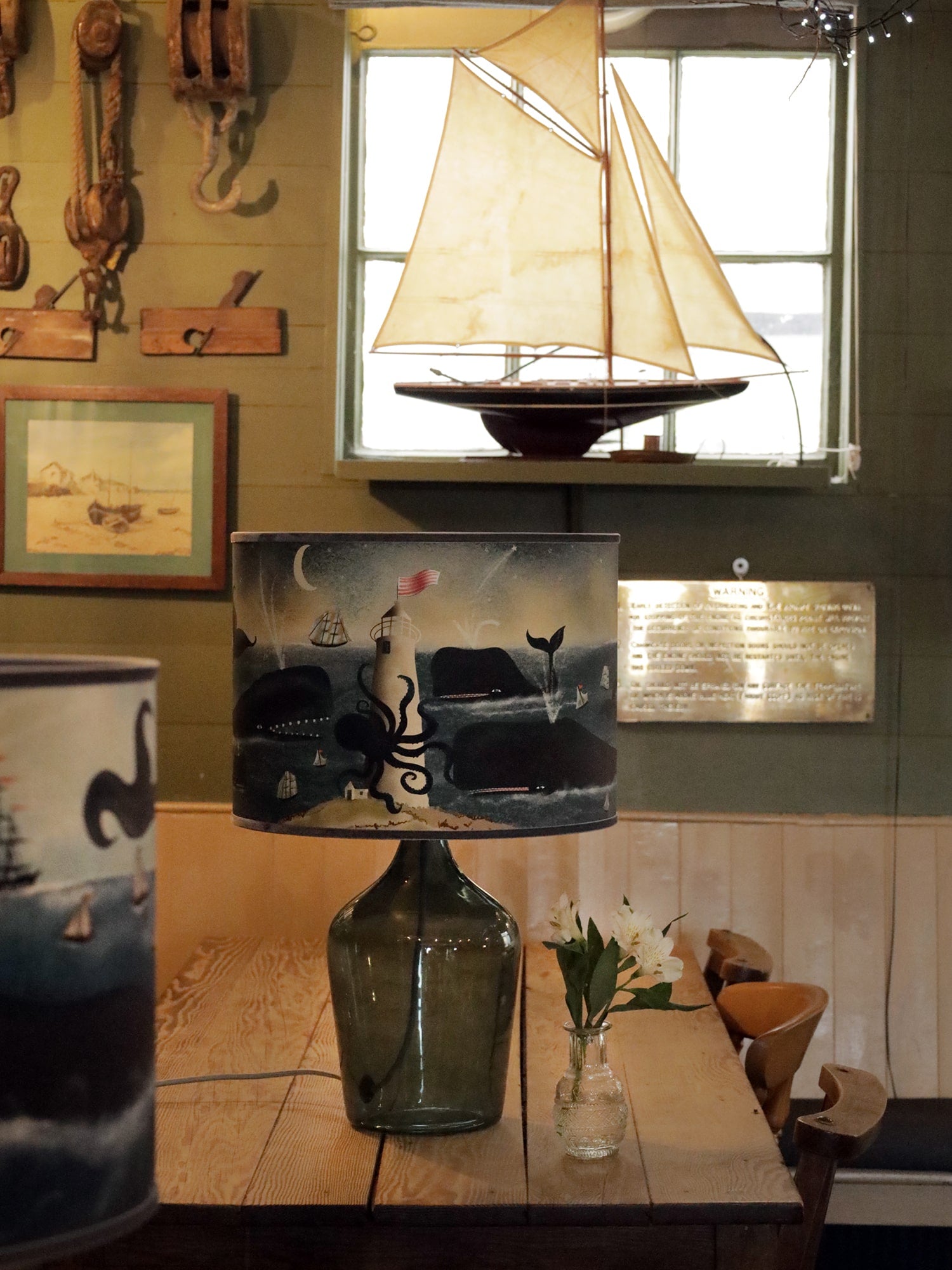 A rustic room with nautical decor featuring a large glass lamp on a wooden table. The lampshade depicts an ocean scene with a lighthouse wrapped by a giant octopus, surrounded by whales and sailboats under the moonlight. A small vase of white flowers sits beside the lamp. In the background, a model sailboat with tan sails is displayed on a windowsill, with vintage maritime tools, such as wooden pulleys and ropes, hanging on the wall. A framed painting of a coastal scene adds to the sea-inspired atmosphere o