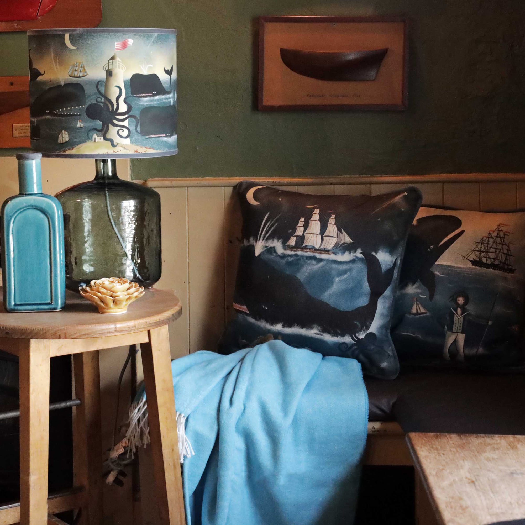 A cozy corner featuring nautical-themed decor, including a wooden side table with a large glass lamp. The lampshade depicts an ocean scene with whales, a lighthouse, an octopus, and sailboats. On the table, a teal ceramic bottle and a decorative ceramic flower add a touch of charm. Beside the table, a bench is adorned with two maritime-themed cushions—one showing a whale and a tall ship under a crescent moon, and the other depicting a captain and a whale. A soft blue blanket is draped over the bench, comple